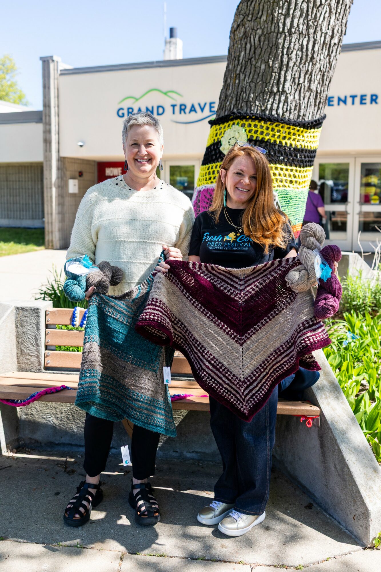 Two women standing outdoors holding colorful knitted blankets, smiling, with a tree and building in the background.