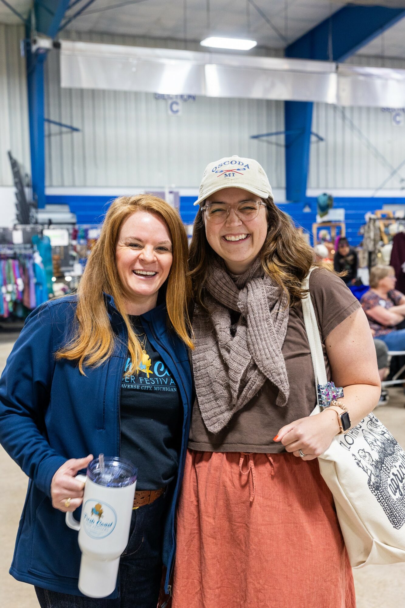 Two women smiling indoors, one holding a water bottle, in a busy event space with booths and people.
