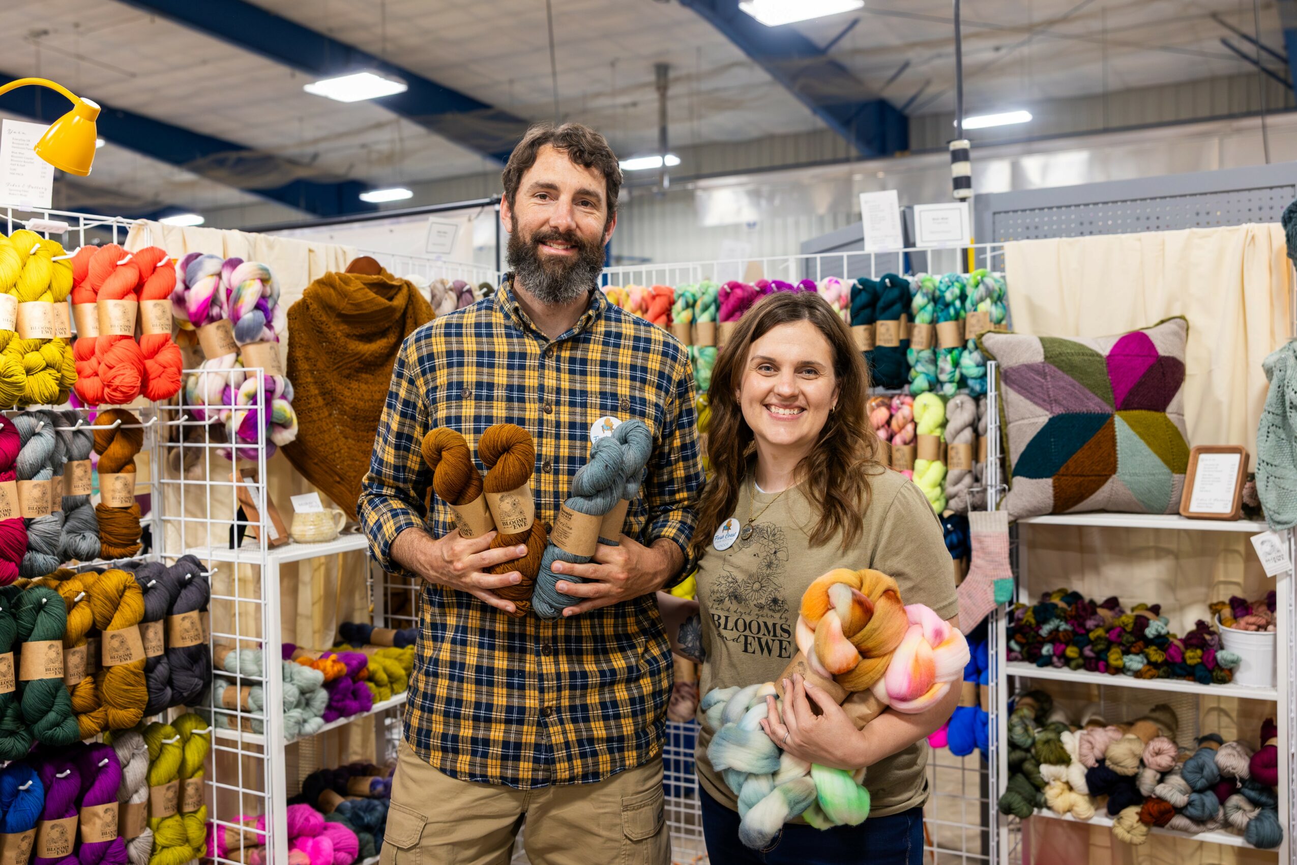 A man and woman stand in a yarn shop holding colorful yarn and stuffed animals, smiling at the camera.