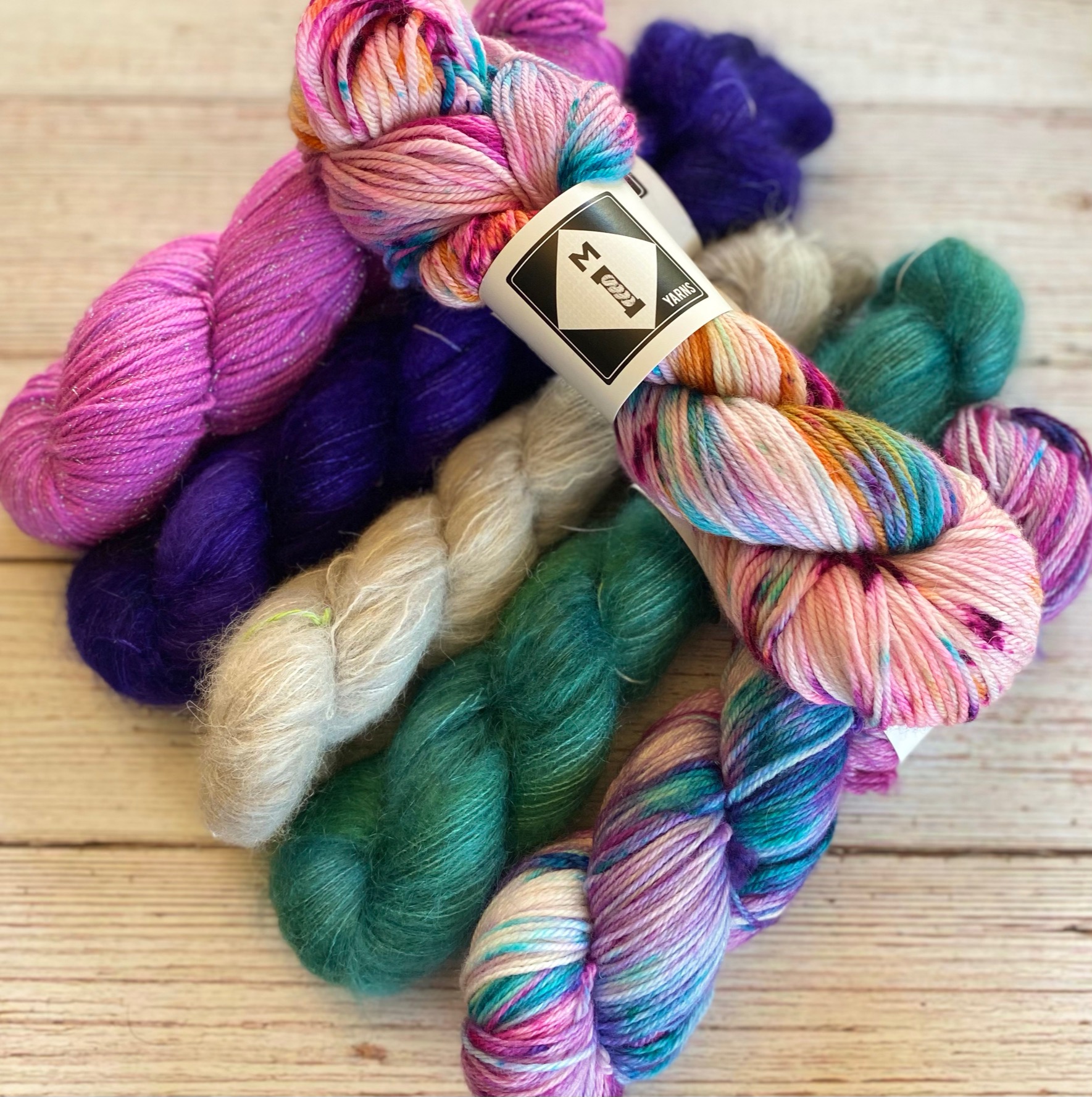 Colorful skeins of yarn in pink, purple, cream, green, and multicolor, arranged on a wooden surface.