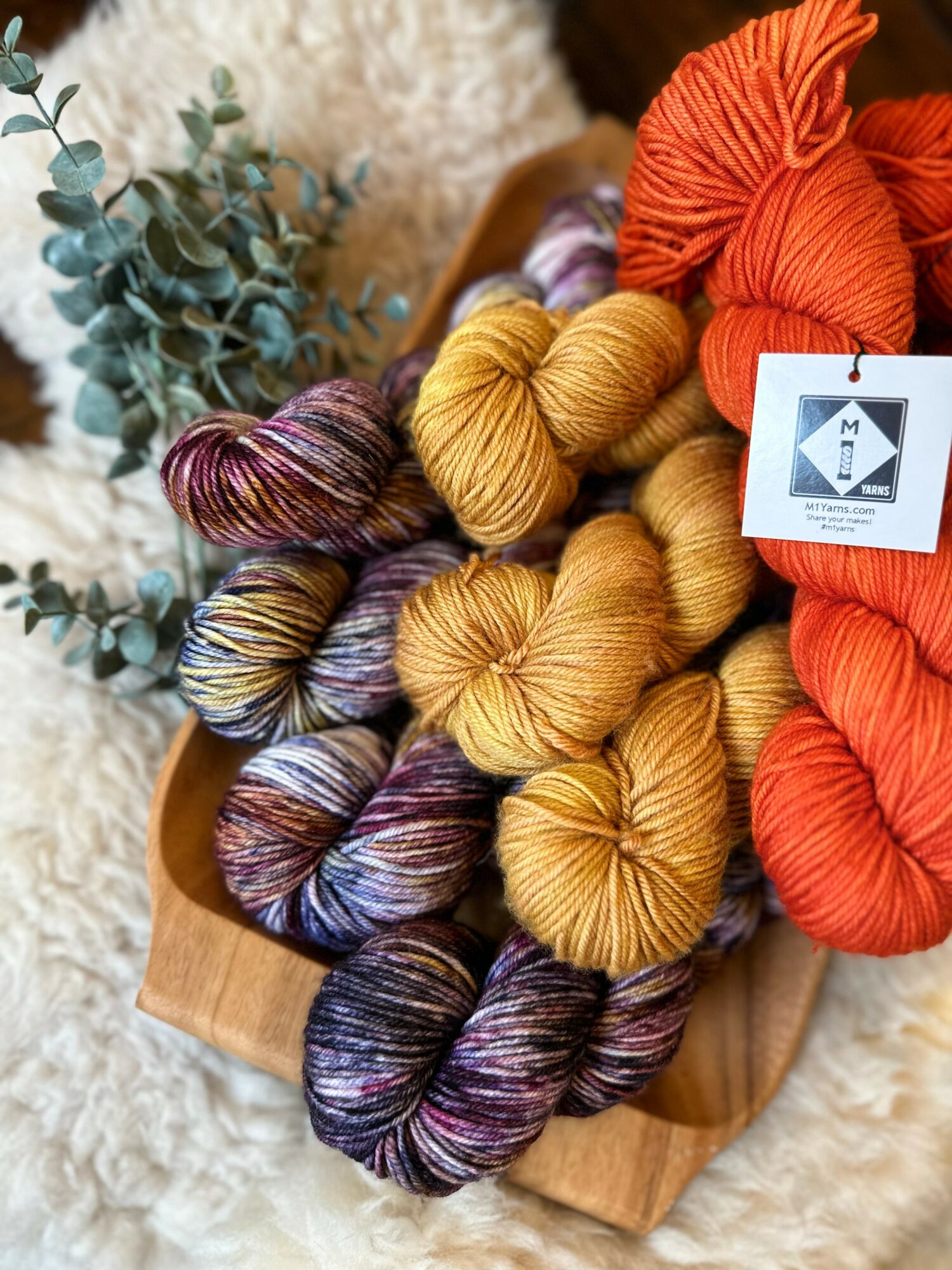 Colorful skeins of yarn in purple, yellow, and orange on a wooden tray with eucalyptus leaves nearby.