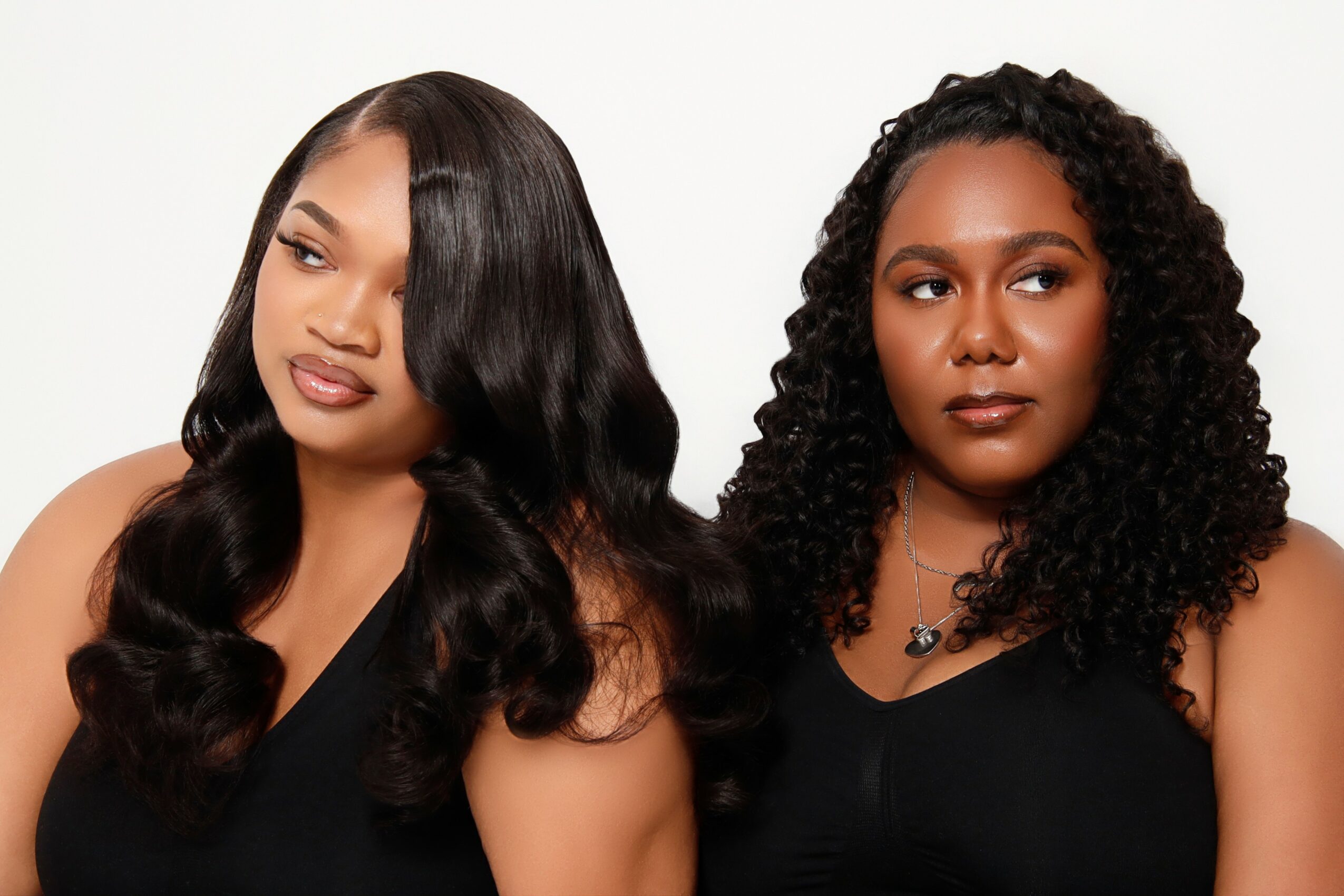 Two women with dark, curly and straight hair, wearing black tops, posing against a white background.