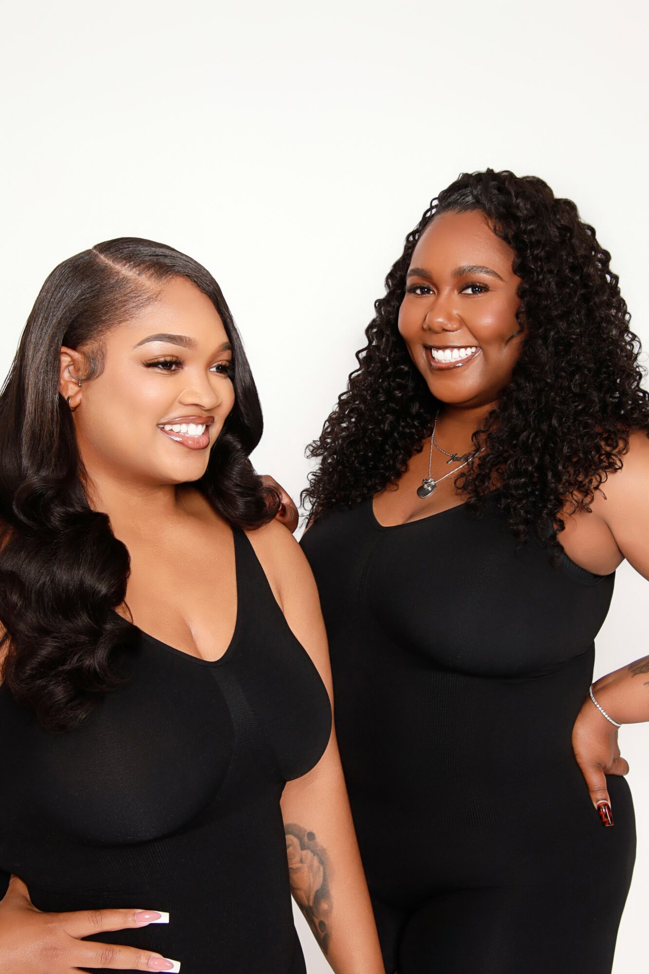 Two women with dark, curly and straight hair smiling, wearing black tops, standing against a white background.