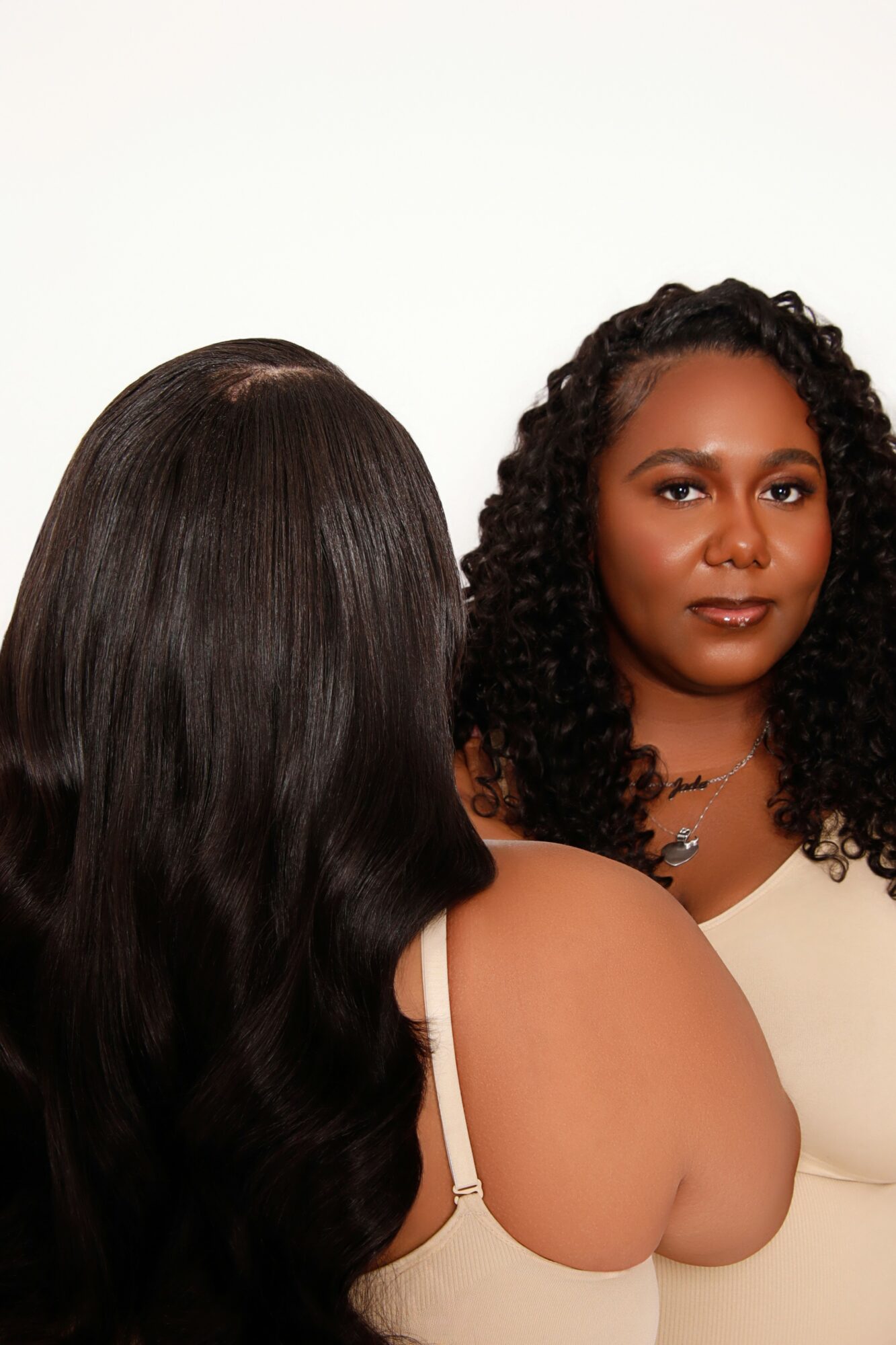 Two women with dark, curly and straight hair, one facing forward and the other with back turned, against a plain white background.