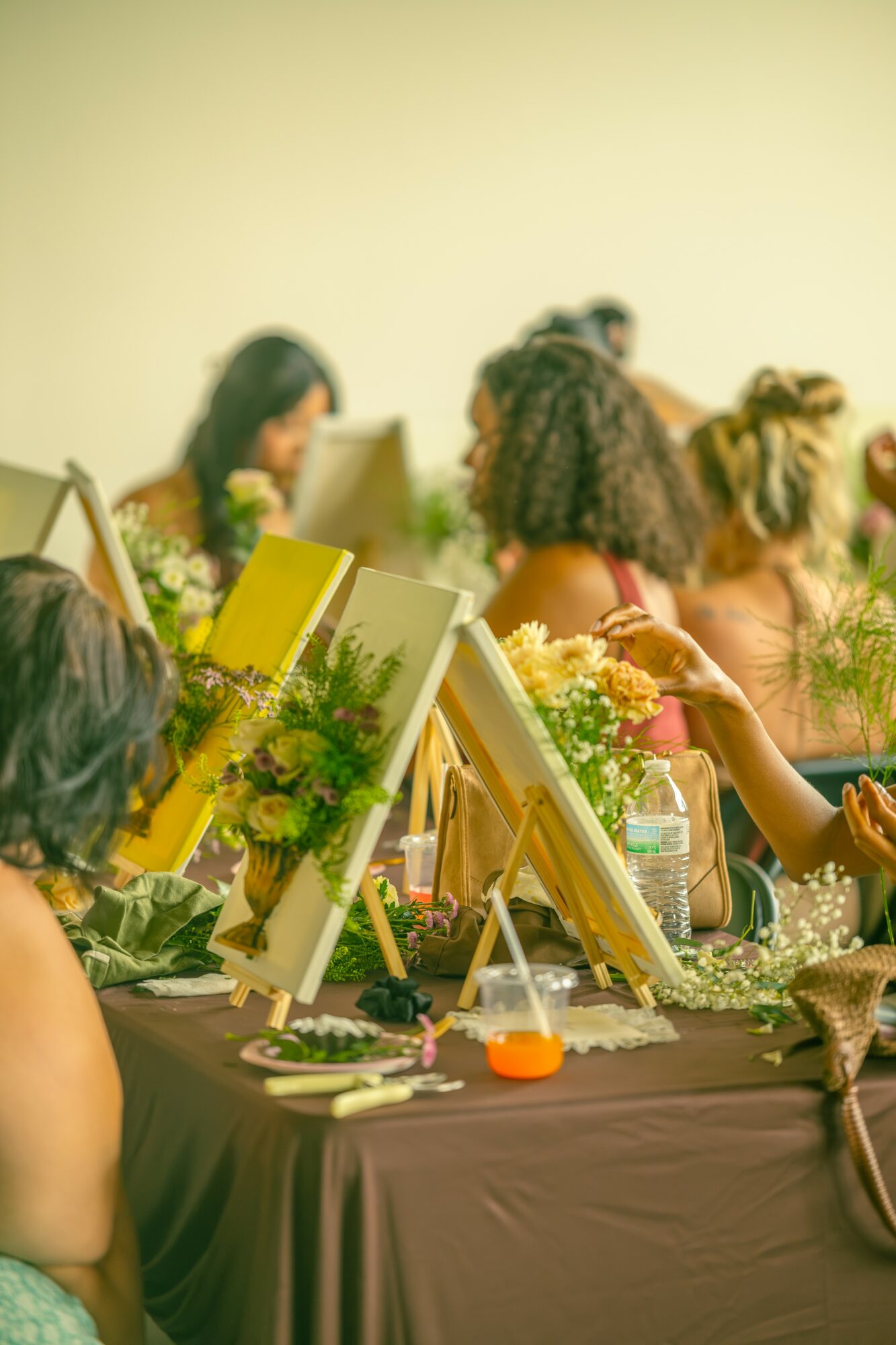 Group of women painting on canvases at a table with art supplies and flowers, in a bright room.