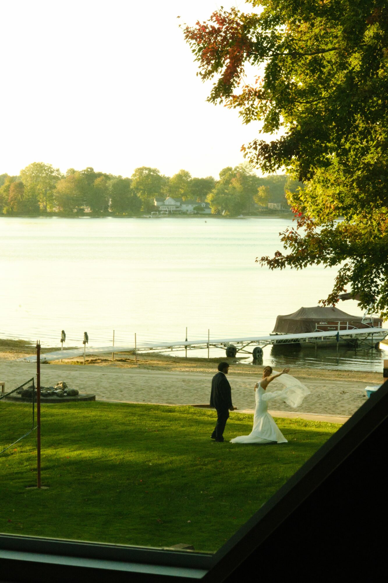 A couple stands on a grassy area near a lake, with trees and a boat in the background, during daytime.