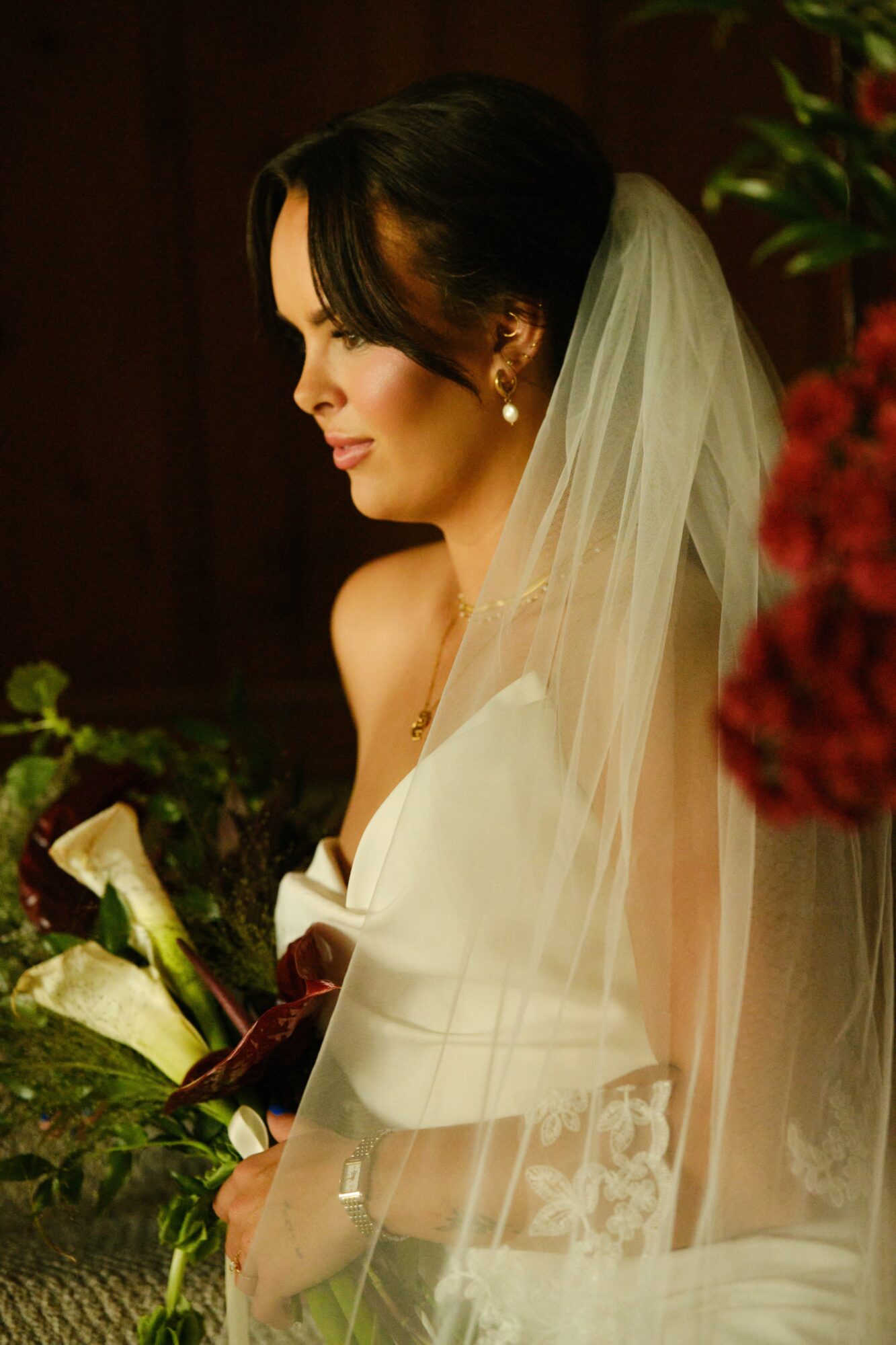 Bride with dark hair in a white dress and veil, holding a bouquet, looking down, indoors with plants and flowers.