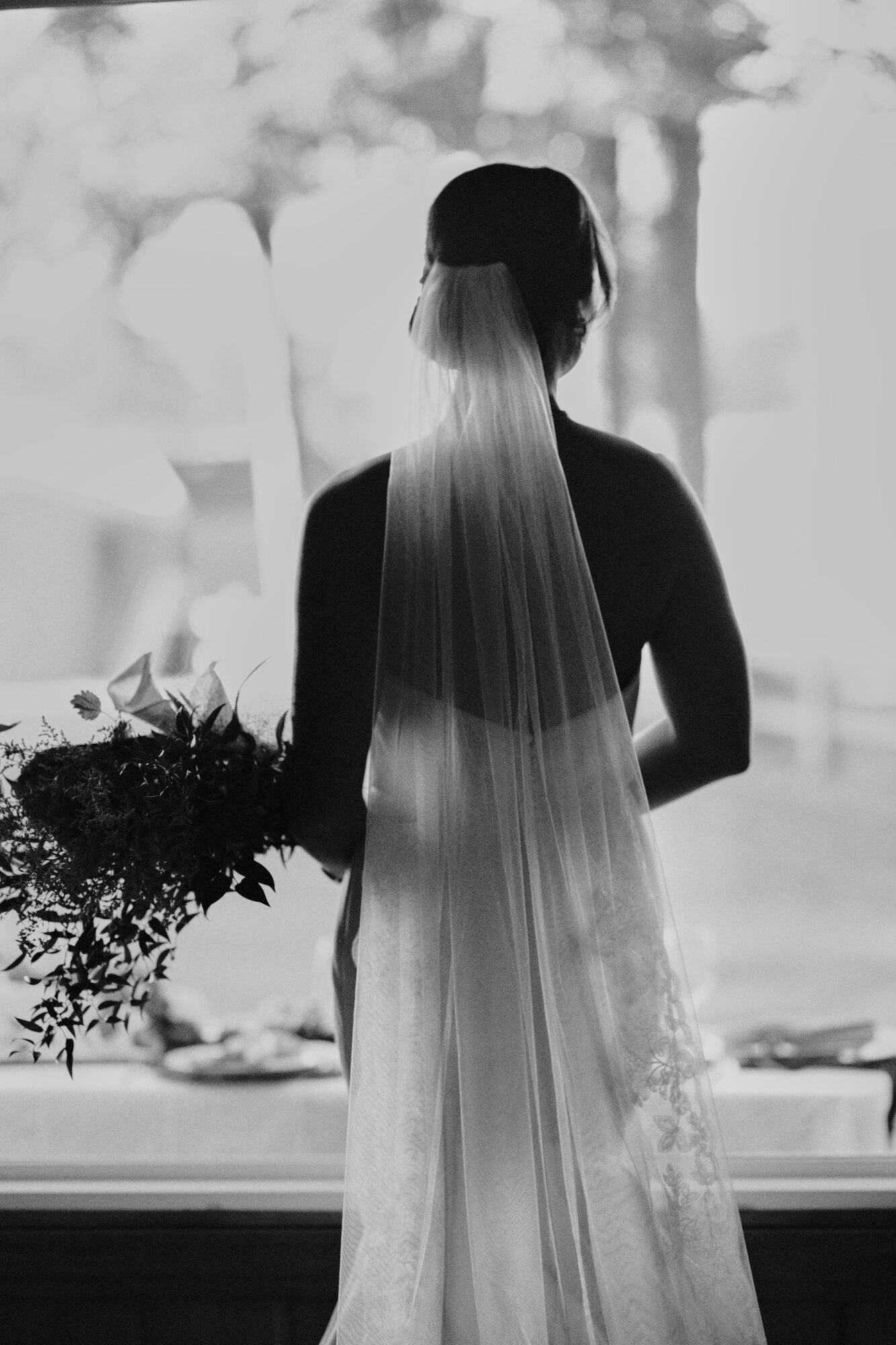 Silhouette of a woman with long, flowing hair, holding a bouquet, standing by a window with trees outside.