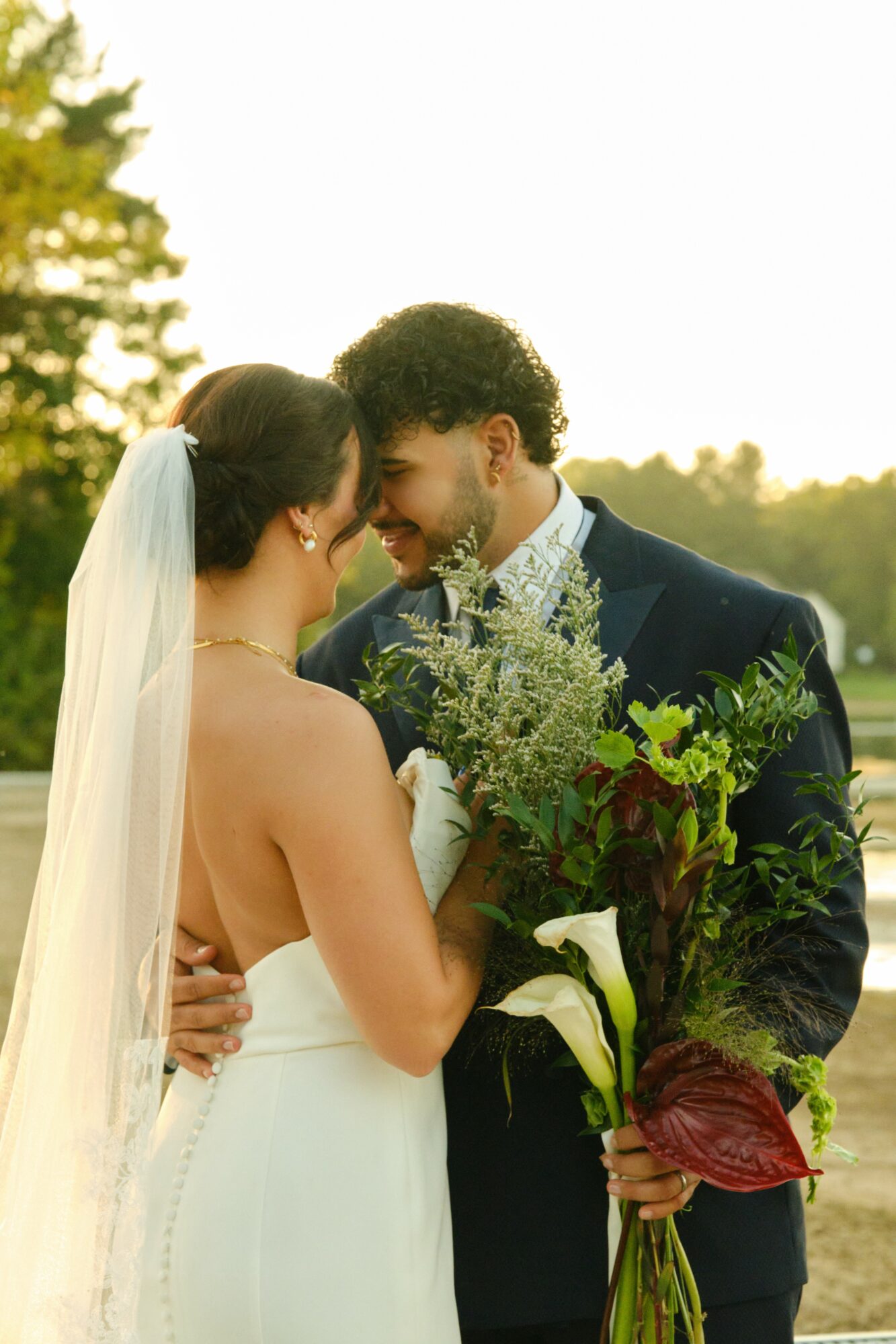 Bride and groom face each other outdoors, holding flowers, smiling, with trees and sky in background.