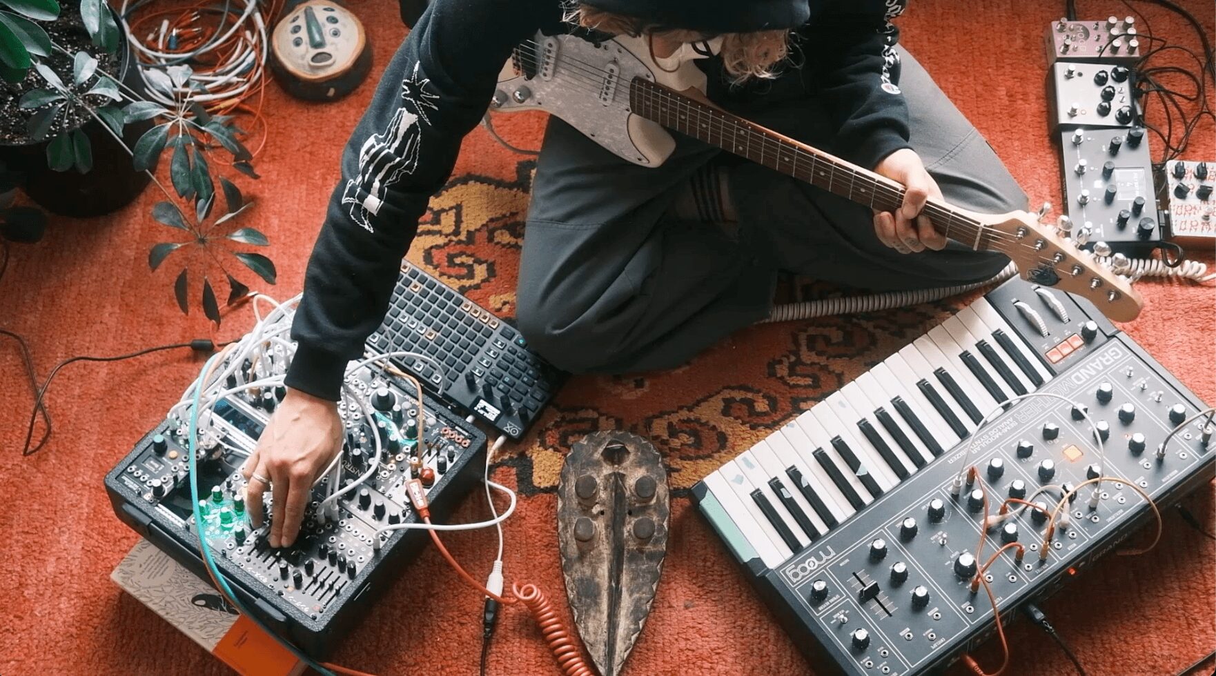 Person sitting on a rug with musical equipment, including a keyboard, synthesizer, and mixing console, adjusting controls.