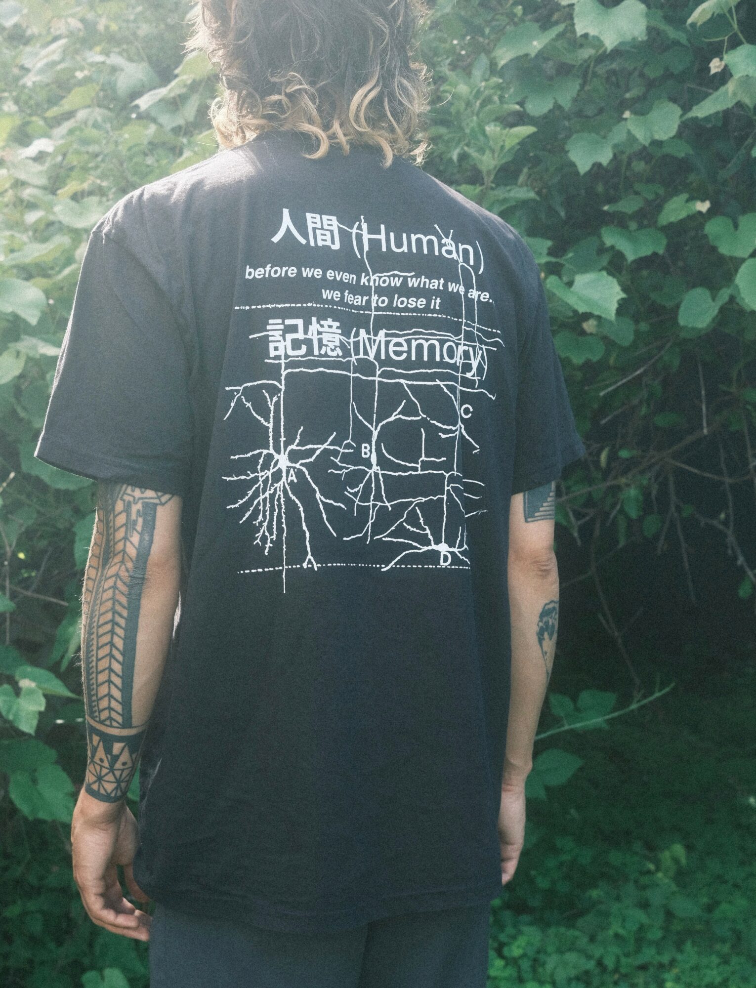 Person with curly hair wearing a black T-shirt with white text and graphics, standing outdoors with green foliage in the background.