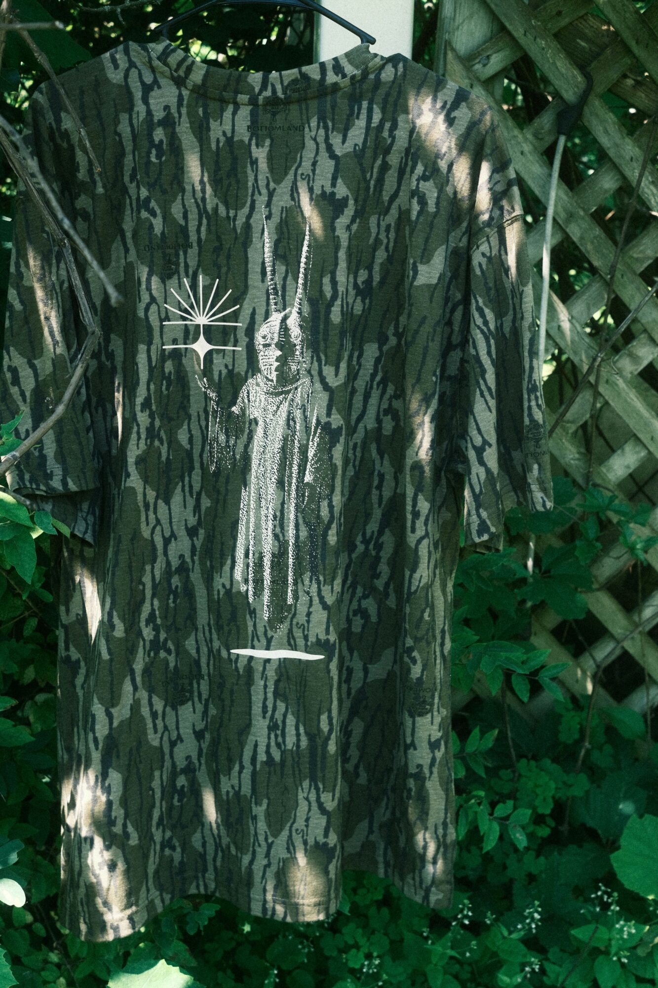 Long dress with a printed image of a woman and a sunburst design, hanging outdoors with greenery and a wooden fence.