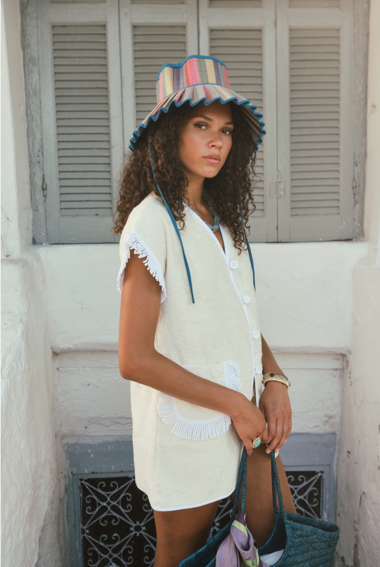 Young woman with curly hair wearing a colorful striped hat and white dress, standing outdoors in front of shutters.