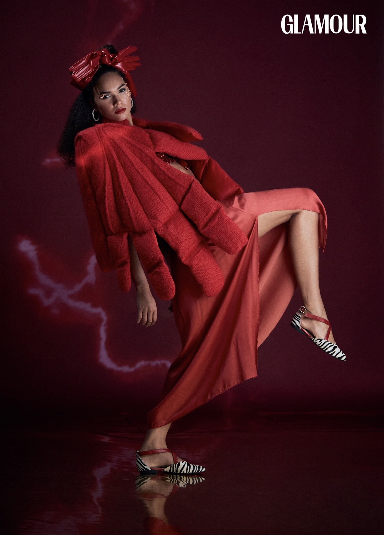 Woman in red dress and jacket poses against a dark background with lightning, wearing striped heels, looking at the camera.