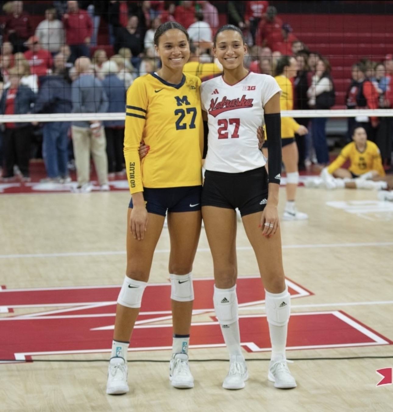 Two female volleyball players stand side by side on the court, smiling, with a crowd in the background.