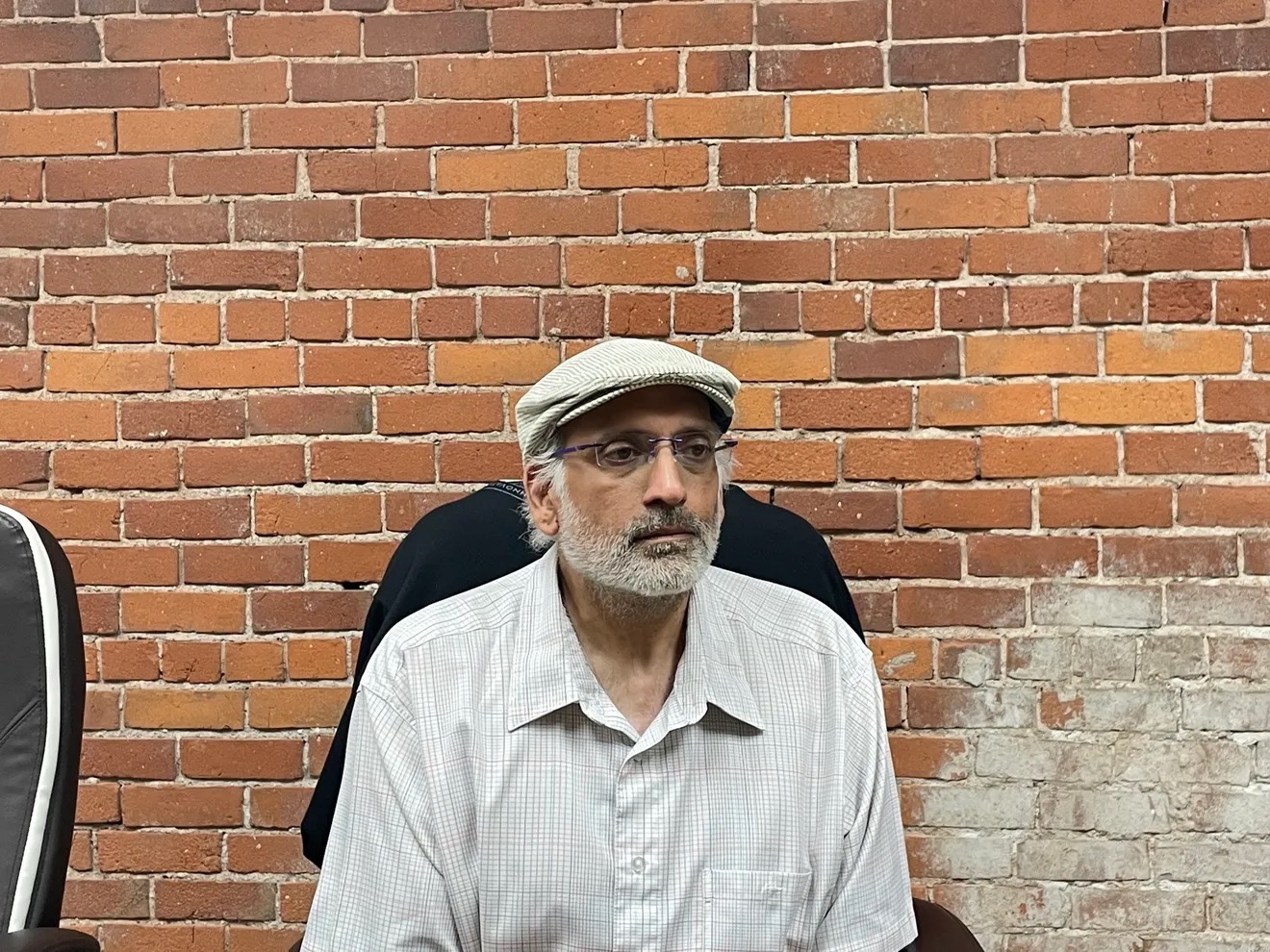 Man with glasses and a cap sitting against a brick wall, wearing a light-colored shirt.