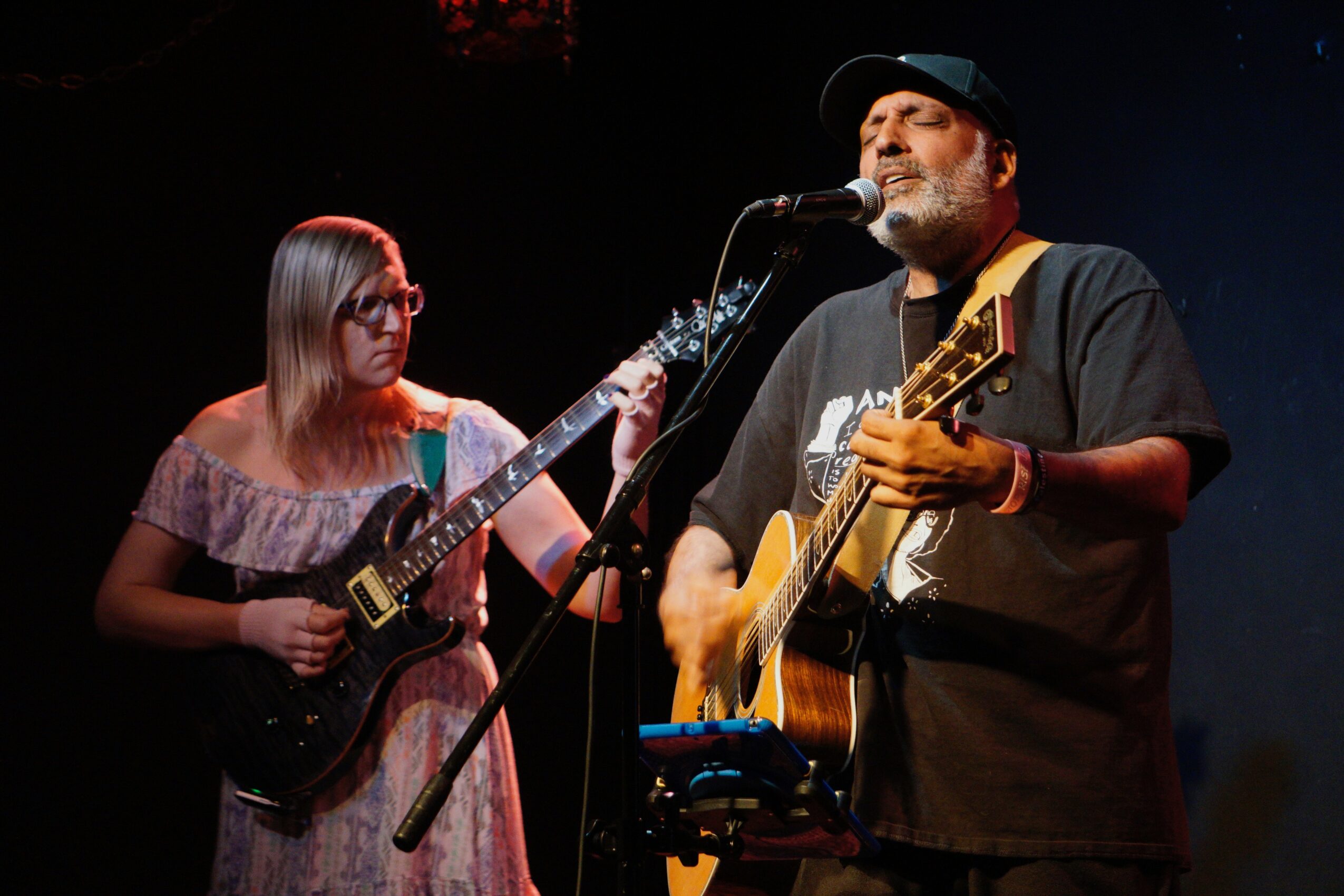 Two musicians perform on stage, one playing an electric guitar and the other singing and playing an acoustic guitar, with dark background.