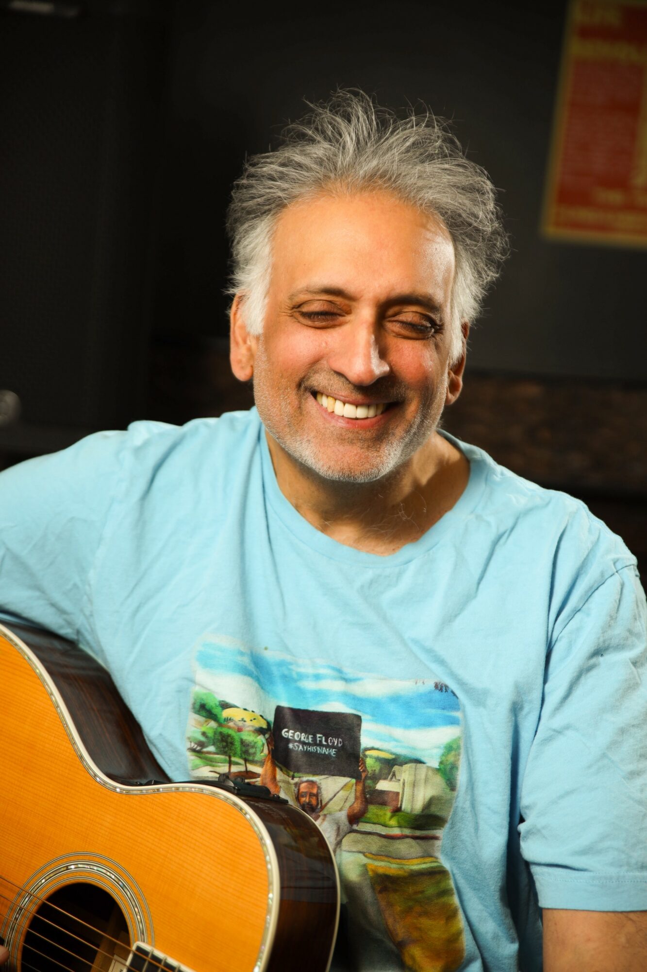 Smiling man with gray hair and beard playing guitar, wearing a light blue T-shirt, in a dimly lit room.