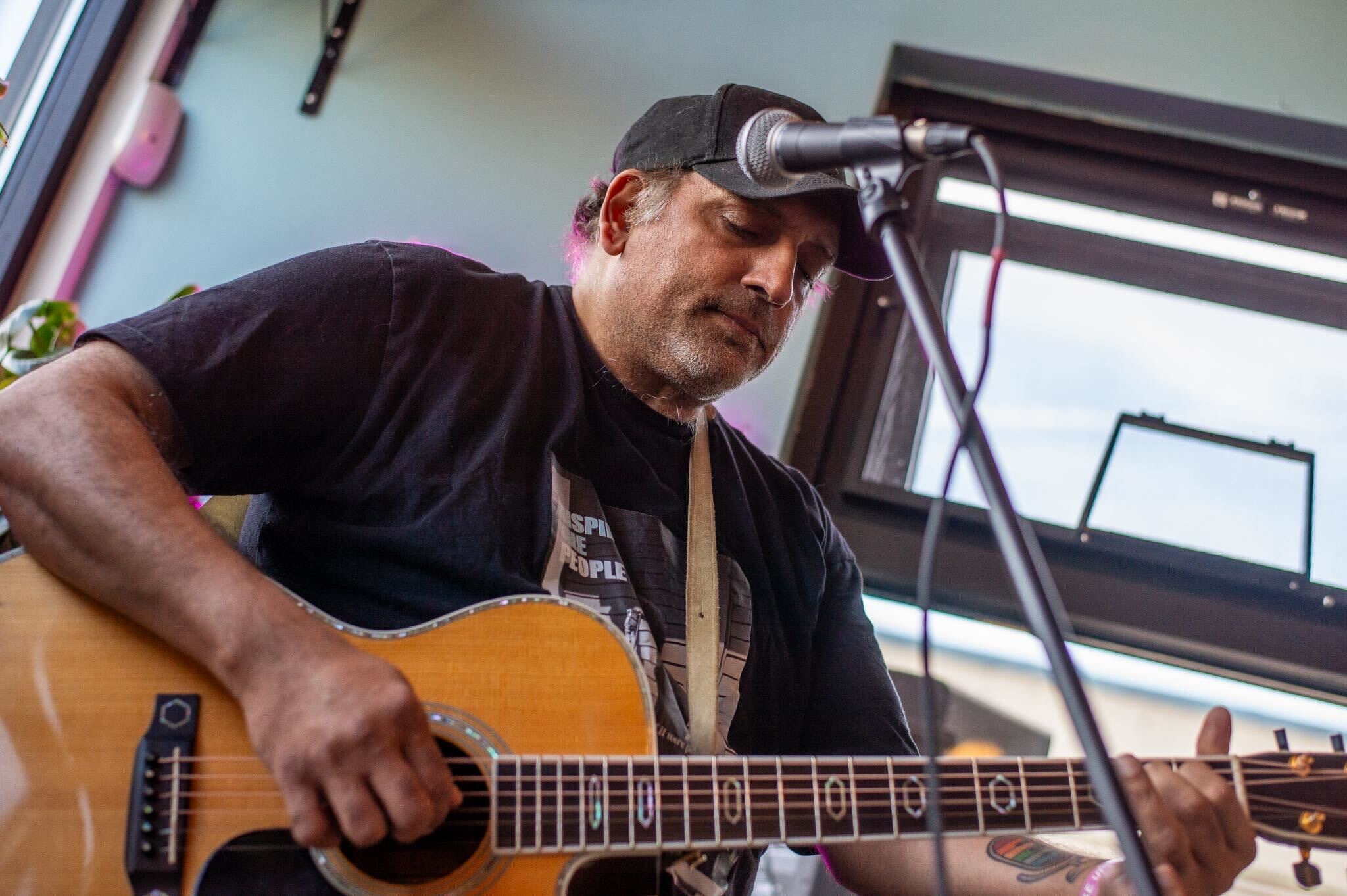 Man playing an acoustic guitar and singing into a microphone indoors, with a window behind him.