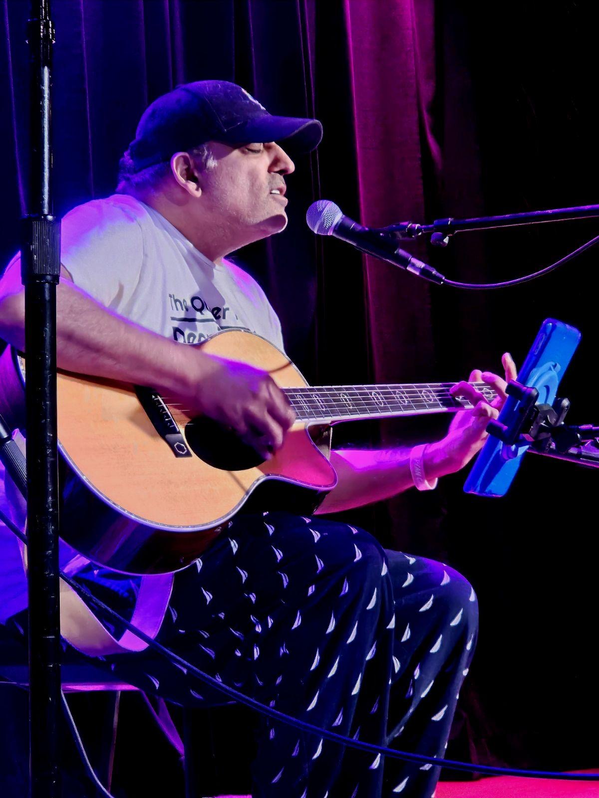 Person playing guitar and singing into a microphone on stage with purple curtains behind.