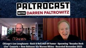 Two men are speaking in a podcast, one with a beard and the other smiling, with bookshelves and curtains in the background.