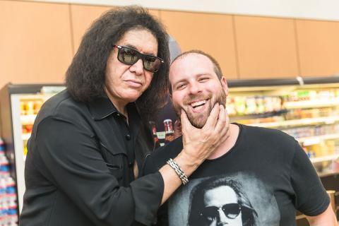 Two men in a store, one with long dark hair and sunglasses, the other smiling with a beard, touching his face.