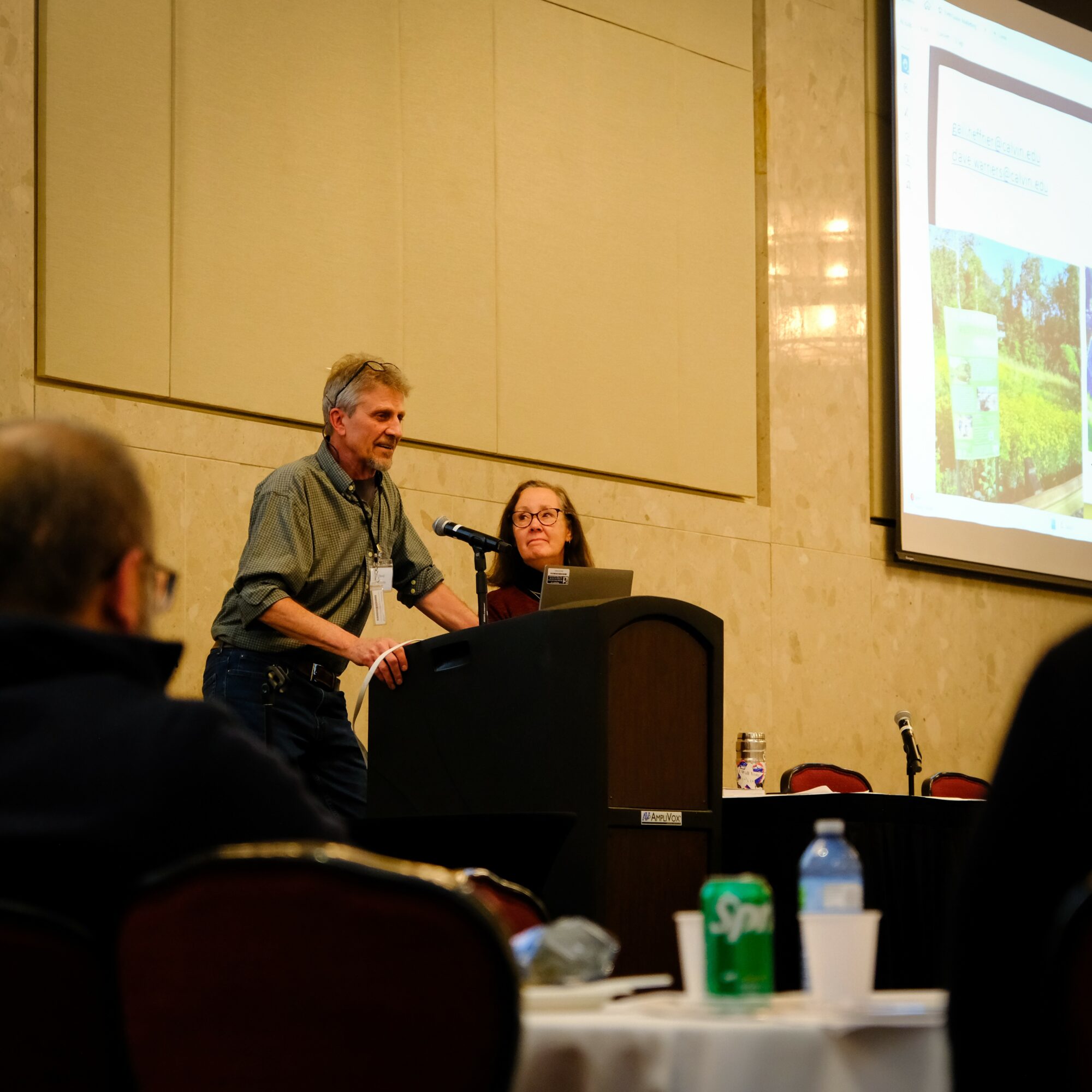 Man speaking at a podium during a presentation, with a woman seated behind him, in a conference room.