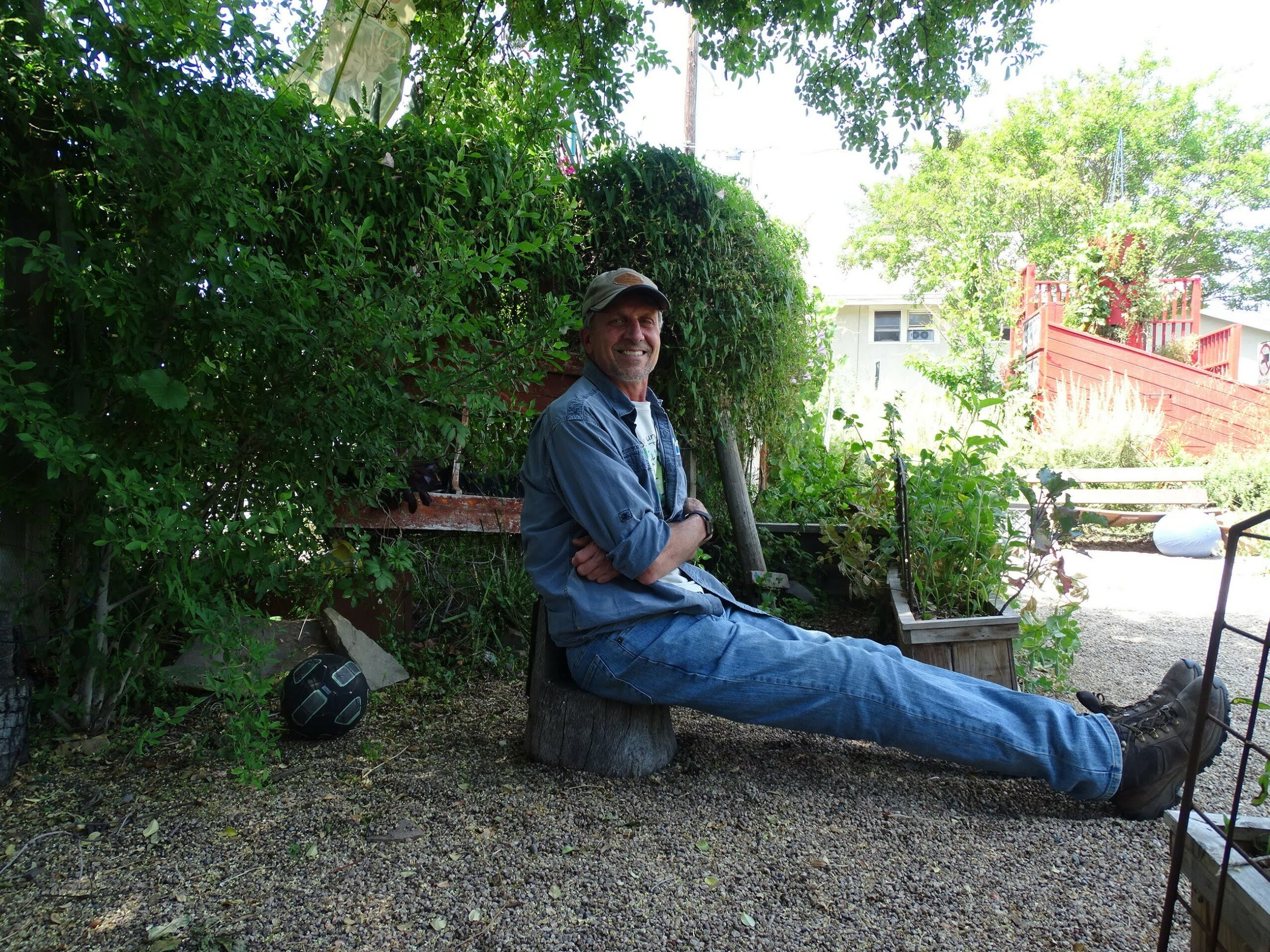 Man sitting on a tree stump outdoors, smiling, with greenery and a building in the background.