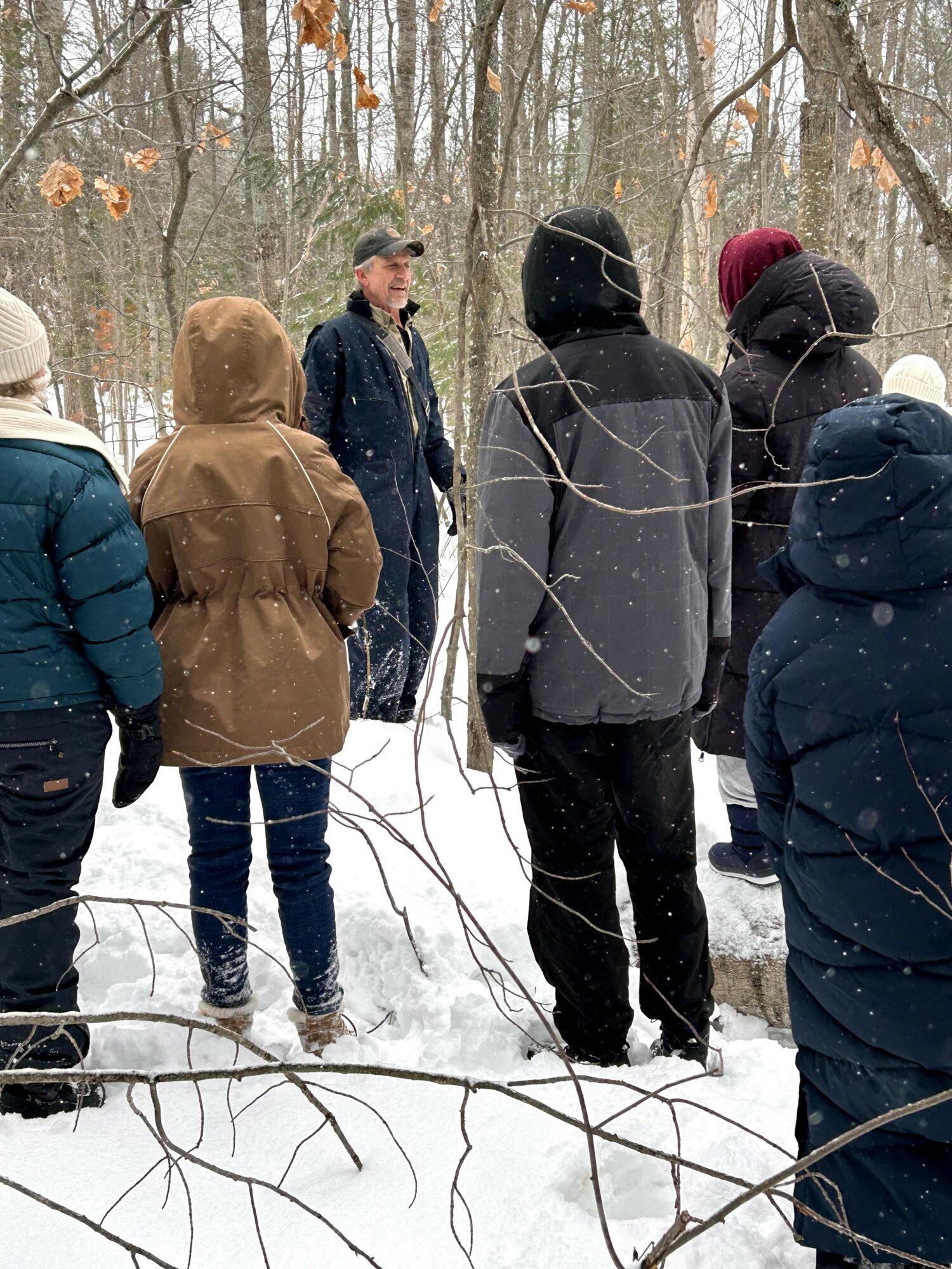 Group of people standing in a snowy forest, listening to a person speaking, wearing winter clothing.
