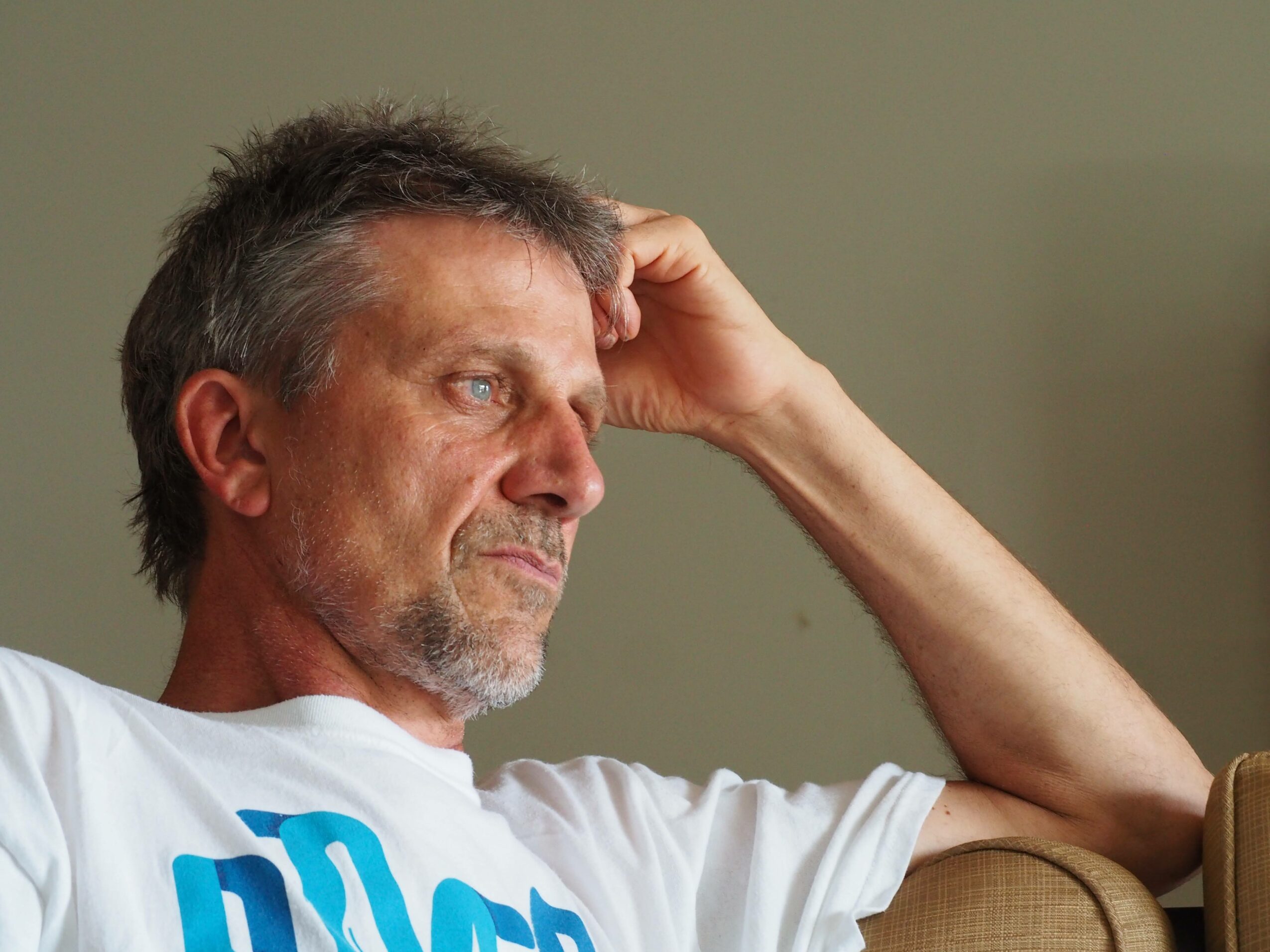 Man with gray hair and beard resting his head on his hand, sitting on a couch, looking thoughtful.