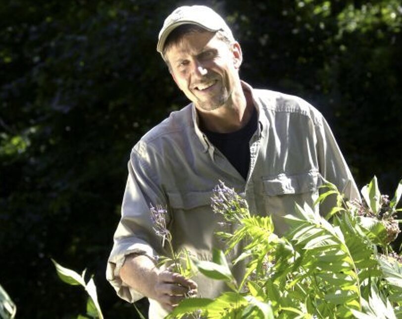 Man outdoors smiling, surrounded by green plants and trees, wearing a light-colored shirt and cap.