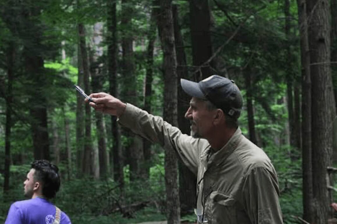 Man in a cap pointing a stick in a forest with trees and greenery, two children nearby, daytime.
