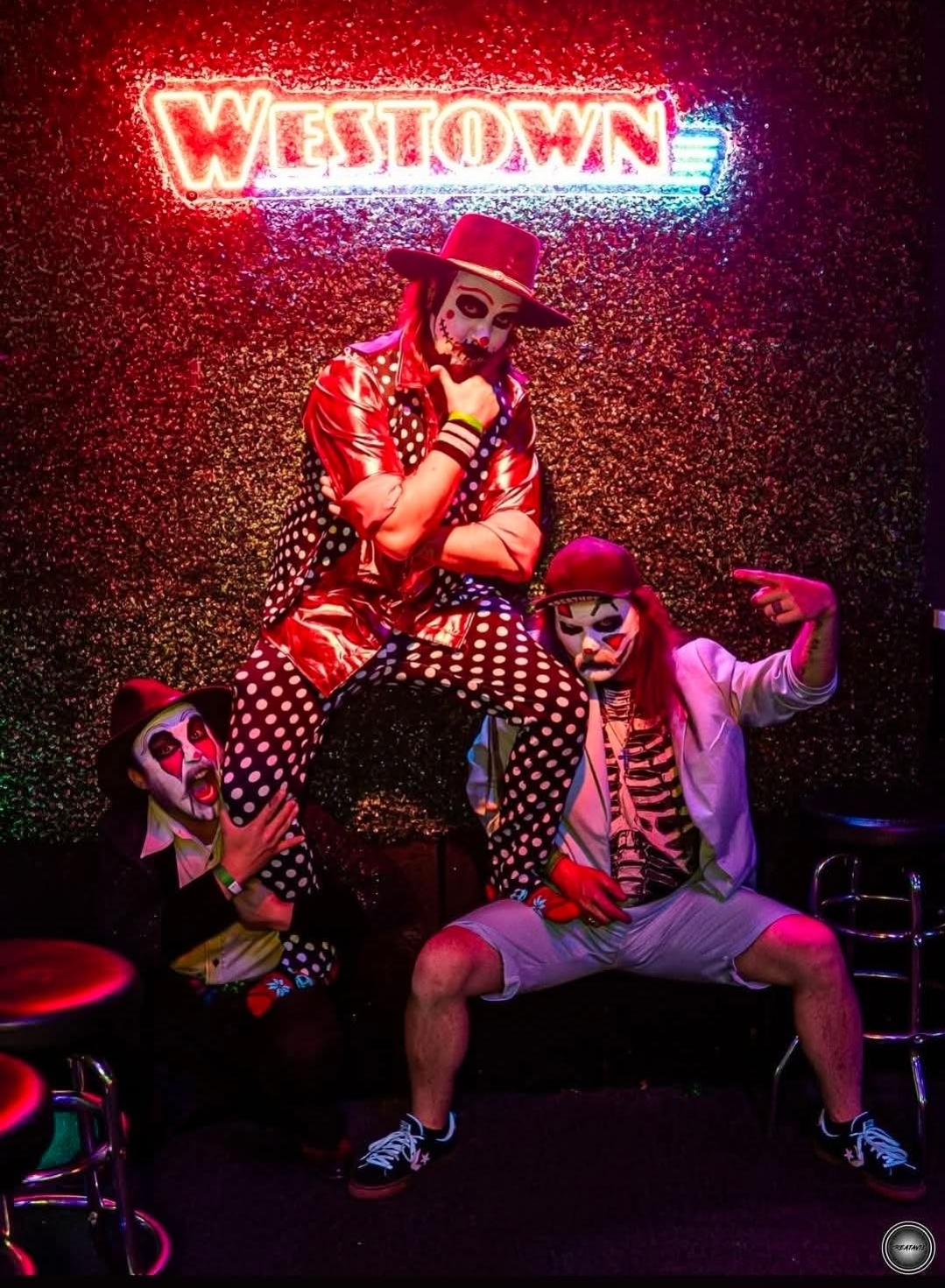 Clown with face paint, wearing a red hat and polka-dot outfit, sitting on a person with clown makeup and striped clothing, in front of a neon sign that says 'Westown'.