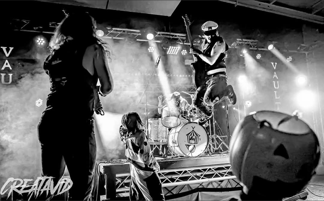Band performing on stage with a drummer, a person with a helmet, and children watching, Halloween decorations visible.