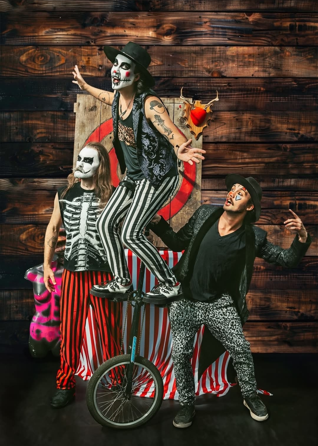 Three performers dressed as skeletons with face paint, one balancing on a unicycle, against a wooden background.