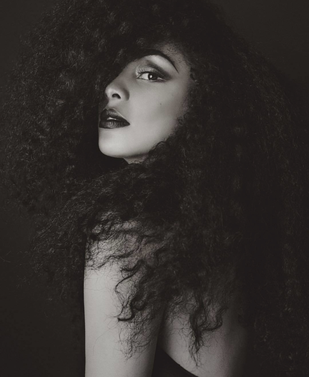 Portrait of a woman with voluminous curly hair, partially turned to the side, with a serious expression, in black and white.