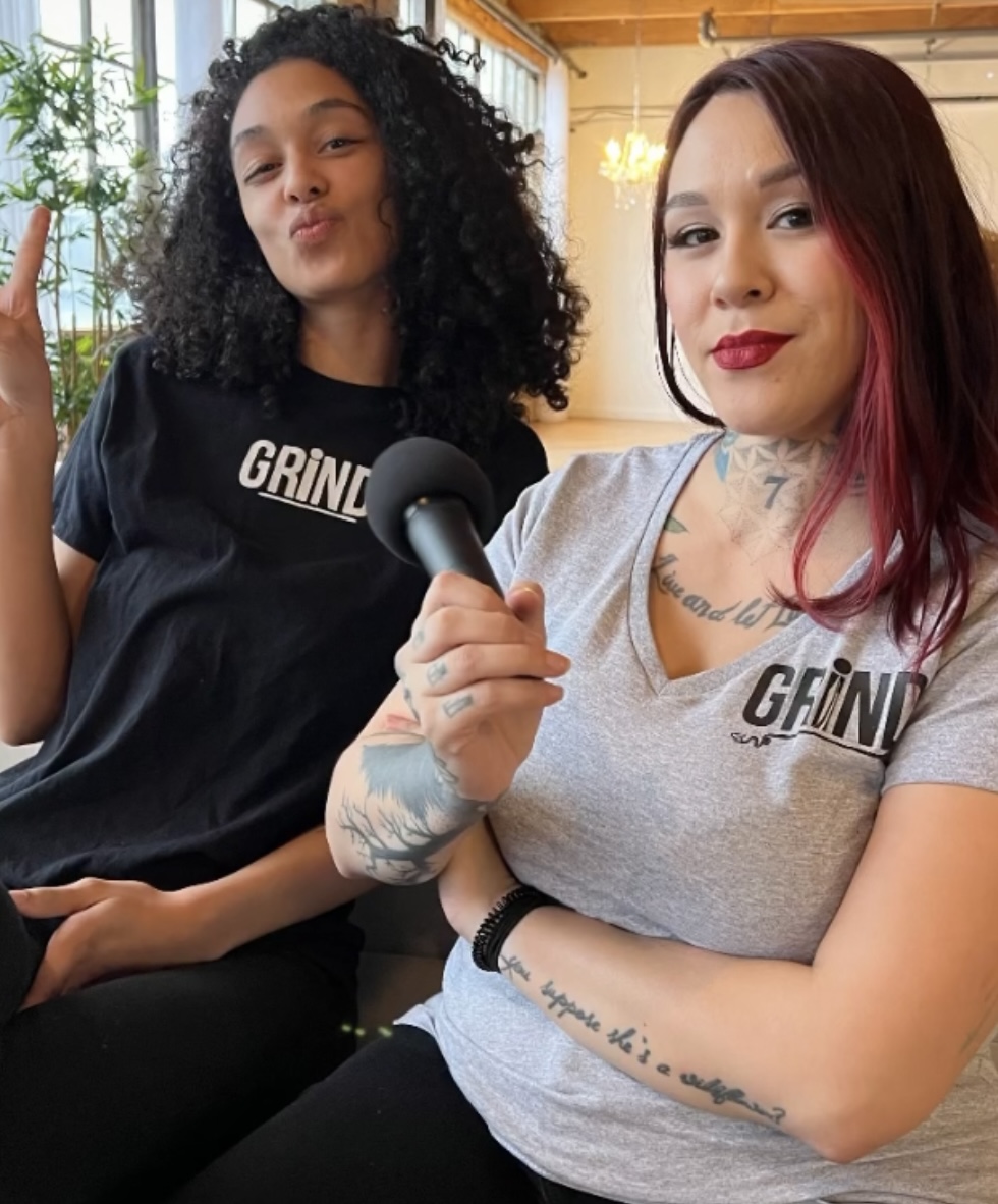 Two women sitting indoors, one holding a microphone, both smiling and wearing gray and black shirts with 'GRIND' text.