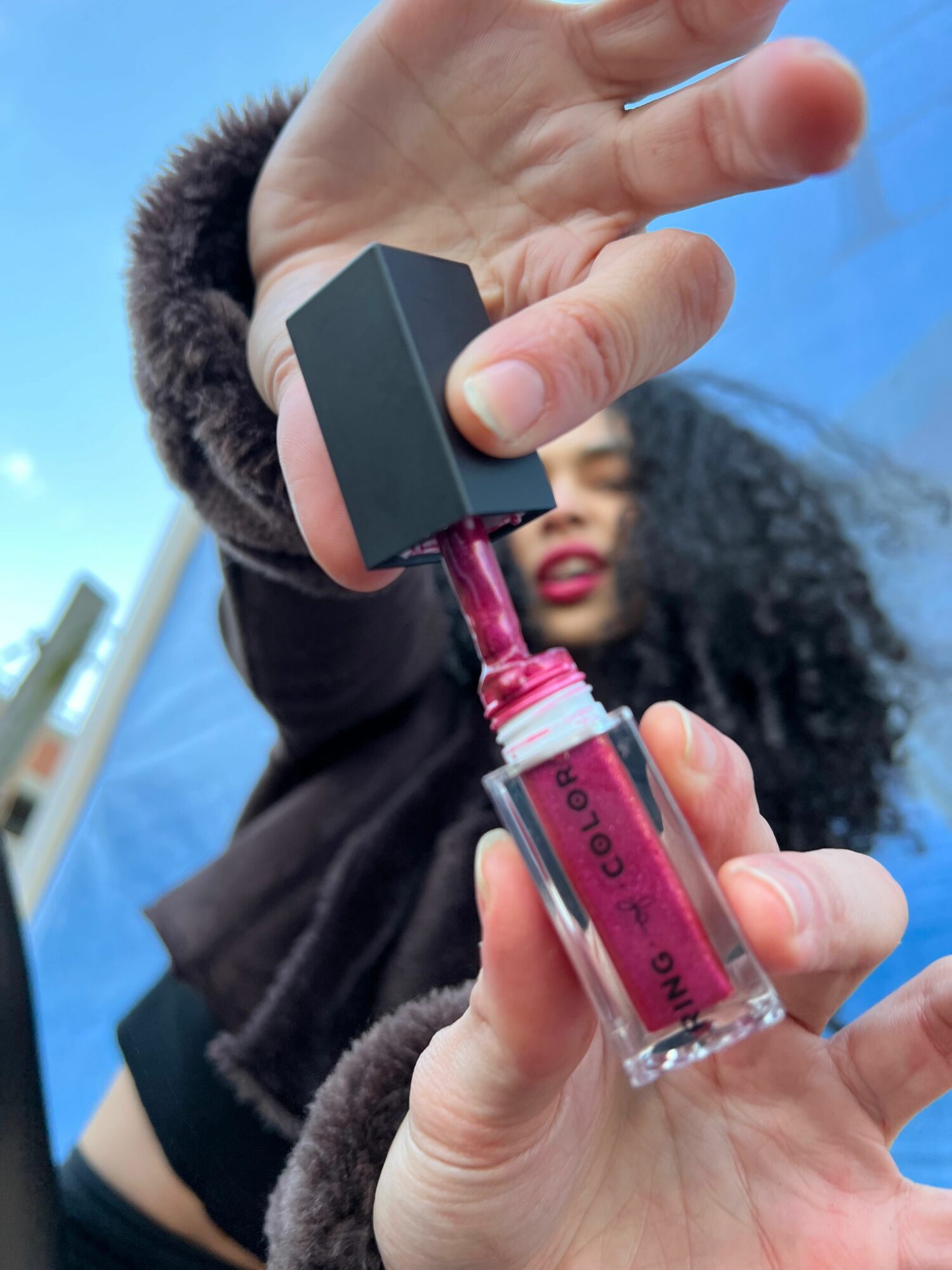 Person holding a small bottle of pink liquid with a black cap, applying it with a dropper, against a blue background.