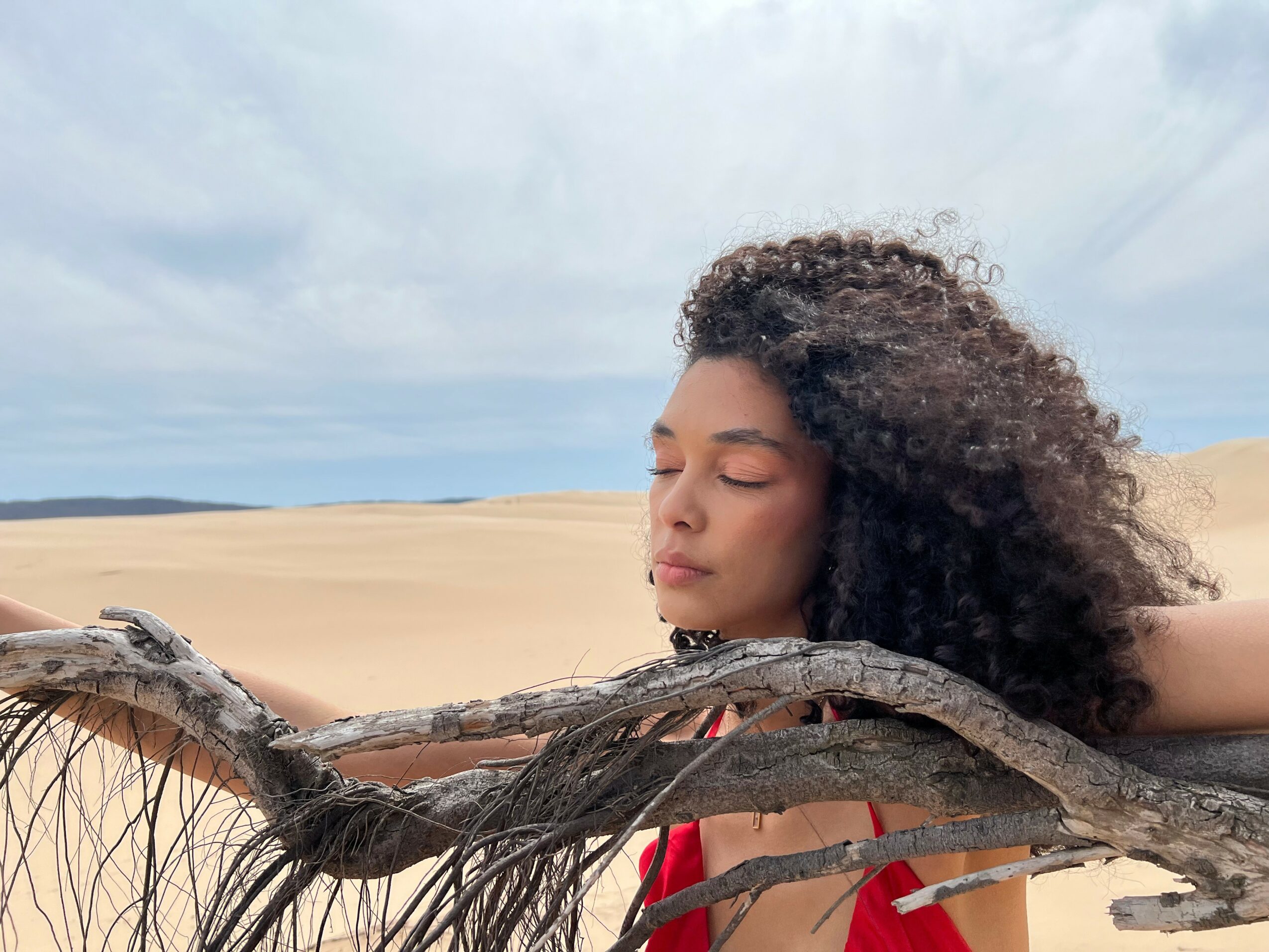 Young woman with curly hair relaxing outdoors with sand dunes and sky in background.