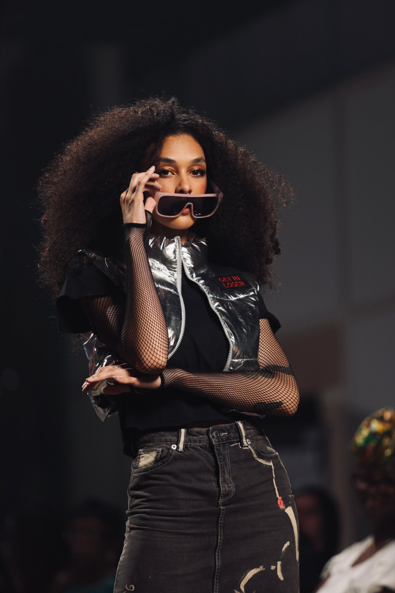 Woman with curly hair wearing sunglasses and a black outfit, standing with arms crossed, in an indoor setting.