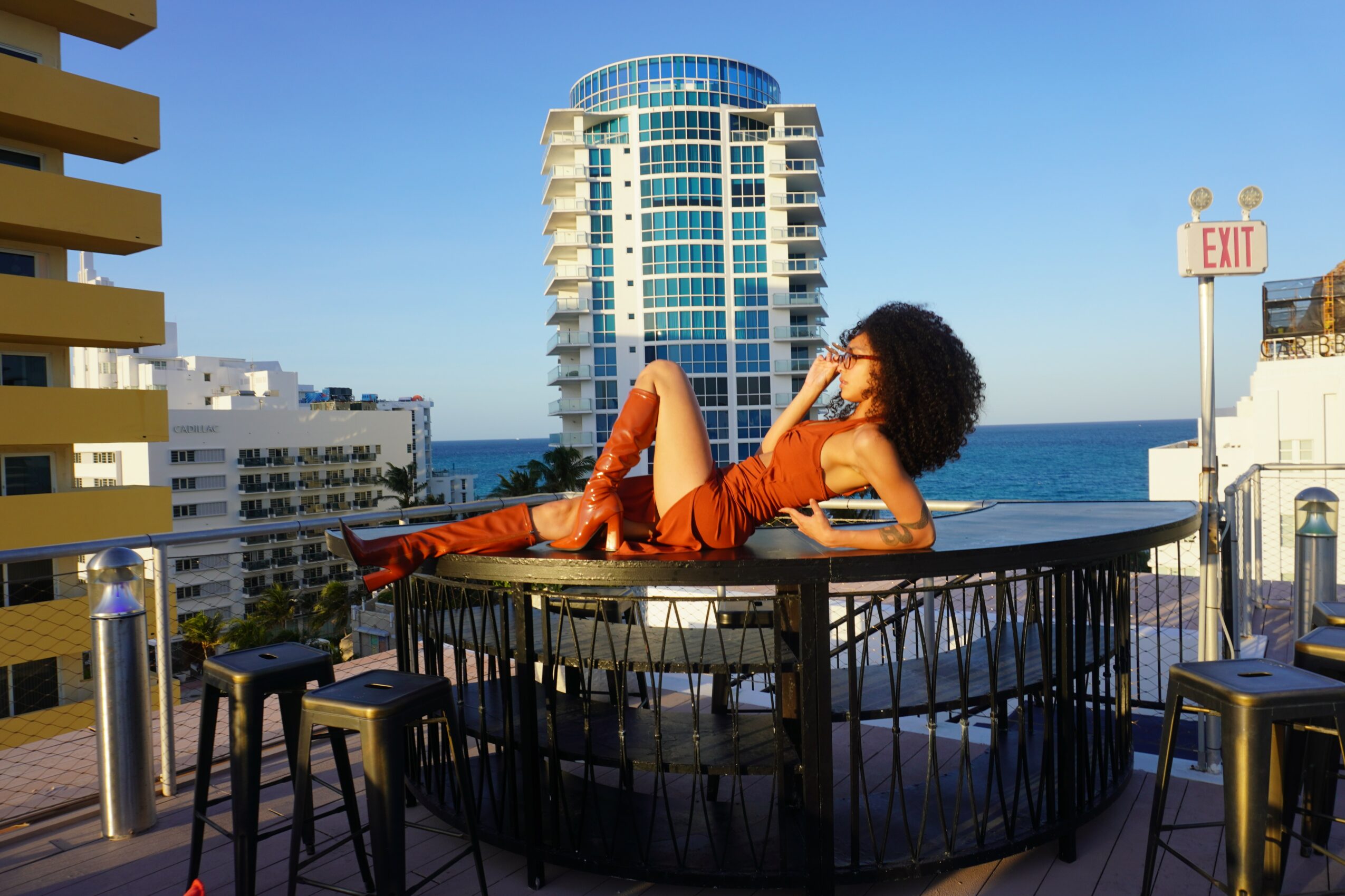 Woman with curly hair in orange outfit reclining on a rooftop table with city and ocean view behind her.