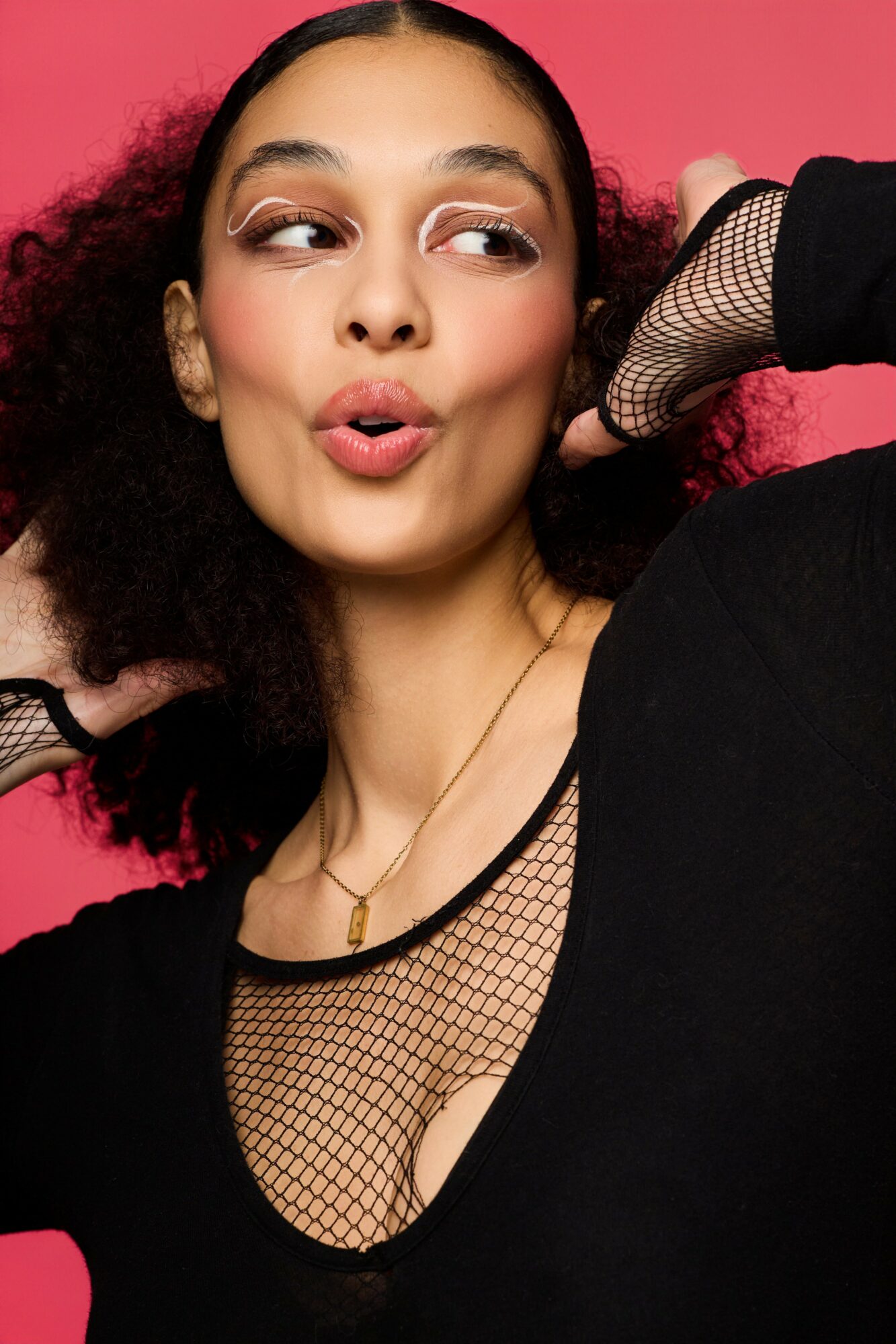 Young woman with curly hair making a playful face, wearing a black fishnet top and a necklace, against a pink background.
