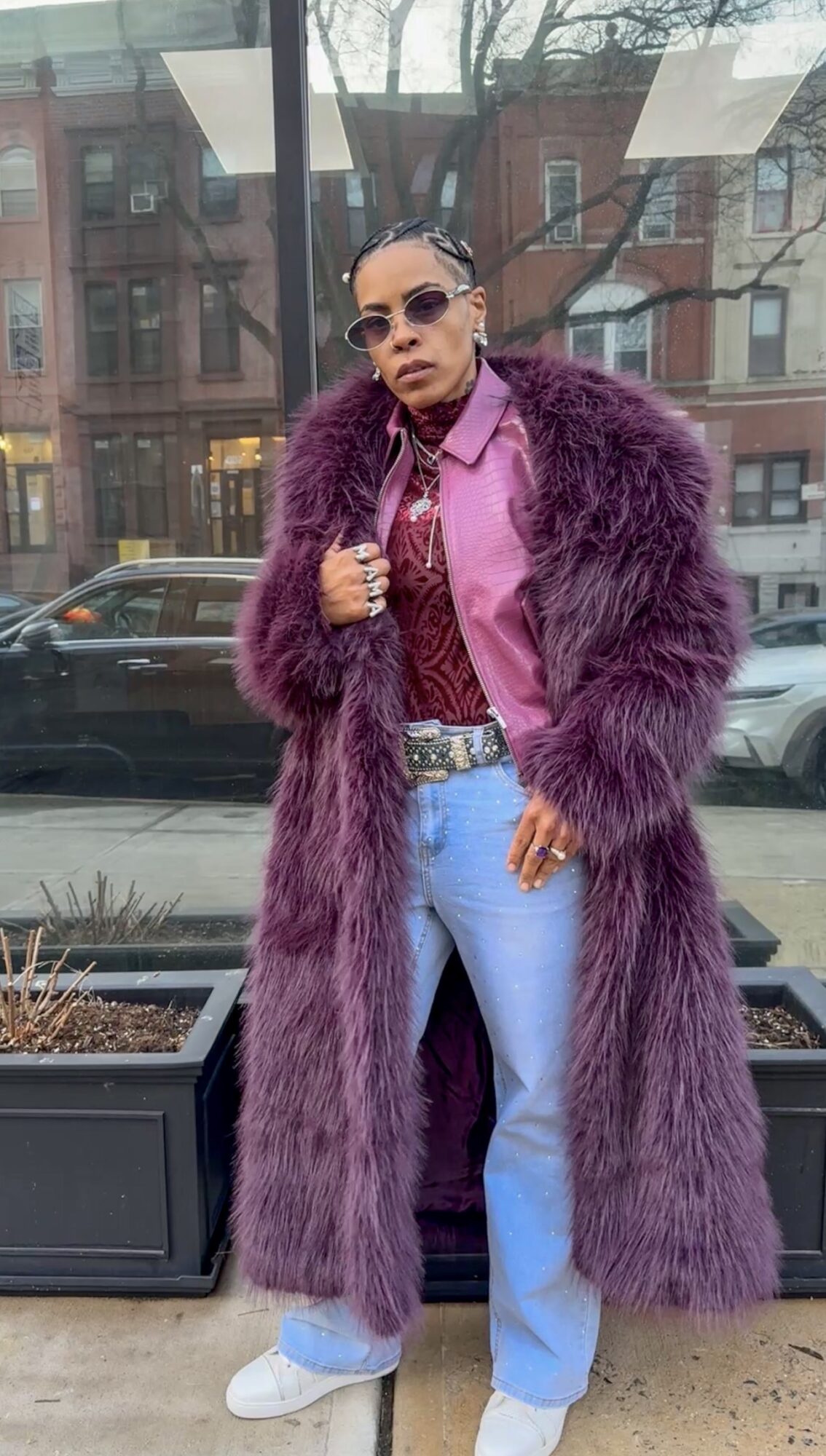 Person wearing sunglasses, a purple fur coat, pink shirt, and light blue jeans, standing outdoors with buildings and cars behind.