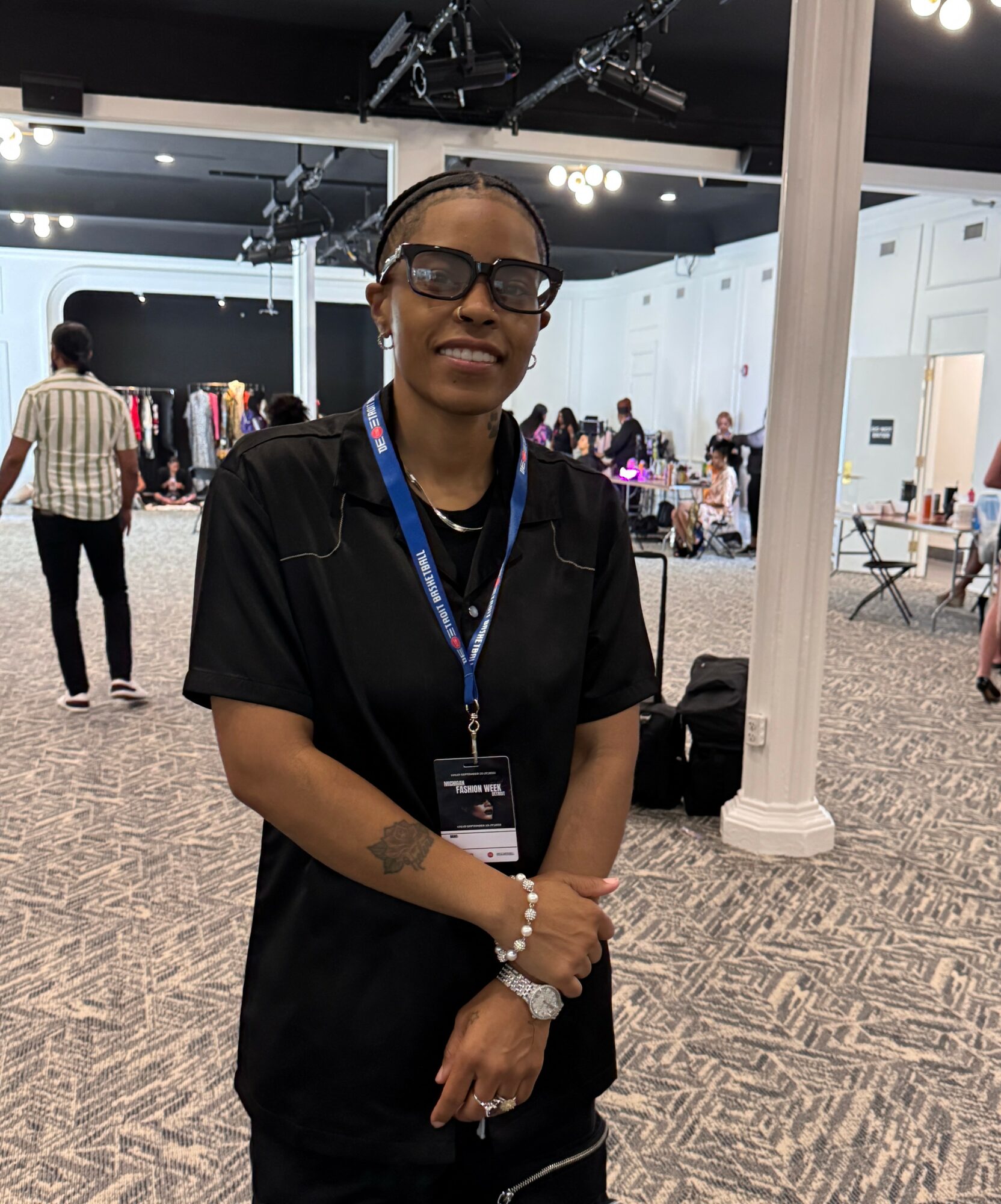 Smiling woman with glasses and a badge, standing in a large indoor event space with people and clothing displays.