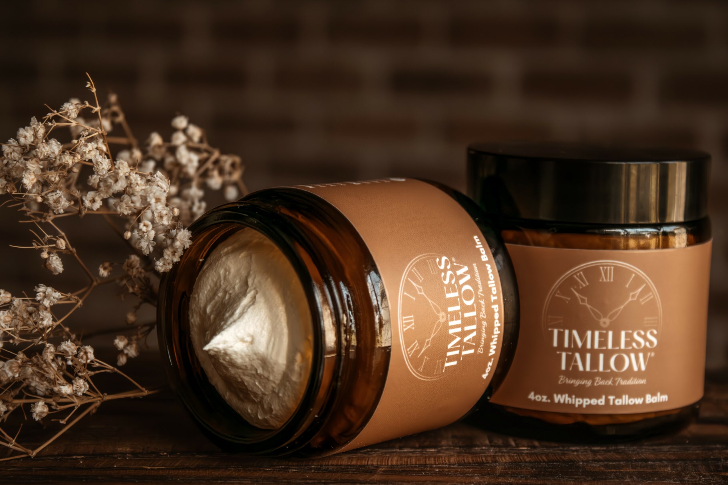 Open jar of Timeless Tallow balm with a black lid, beside dried flowers, on a dark background.