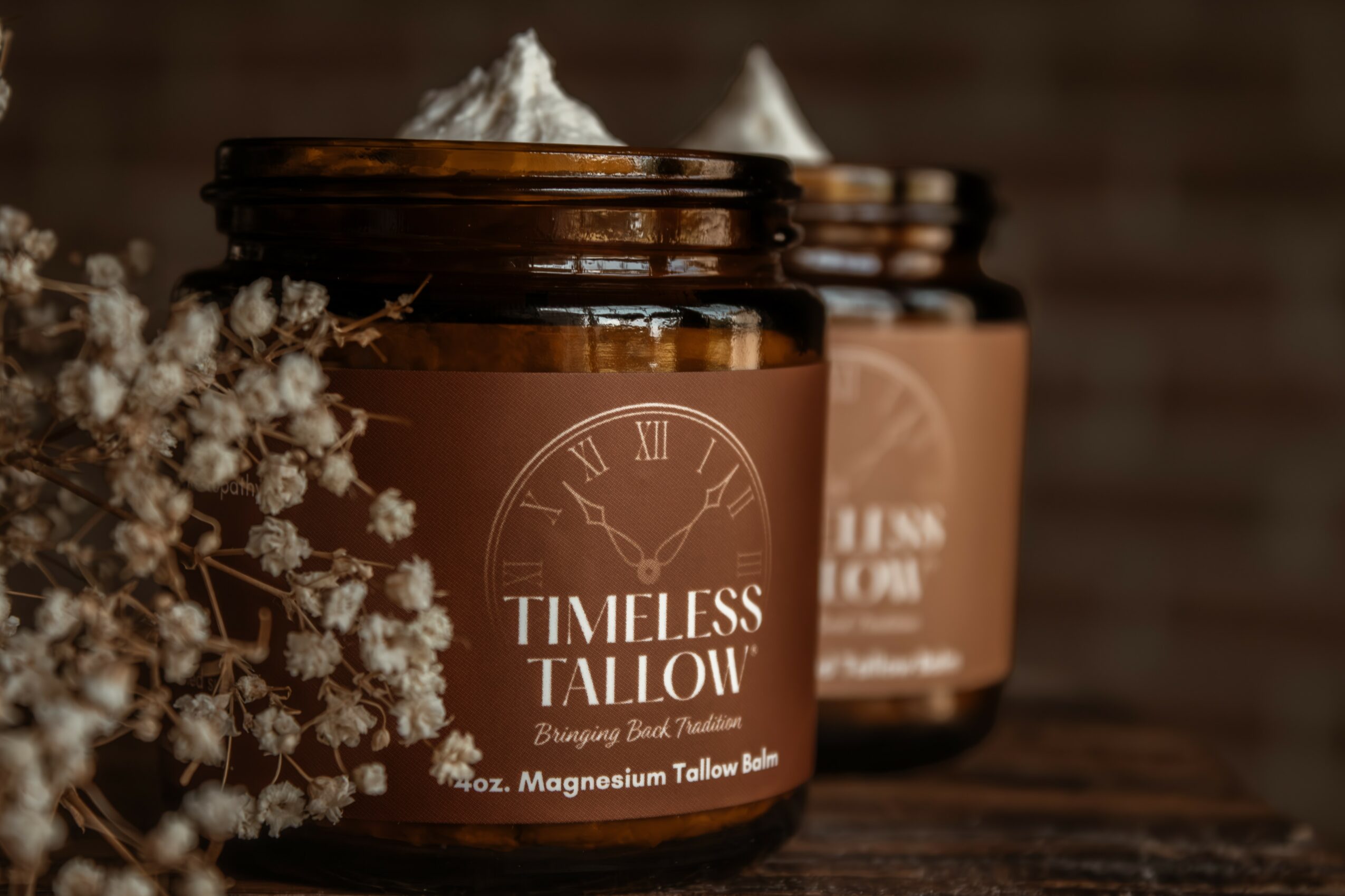 Two jars of magnesium tallow balm with a flower arrangement beside them, on a wooden surface, against a dark background.