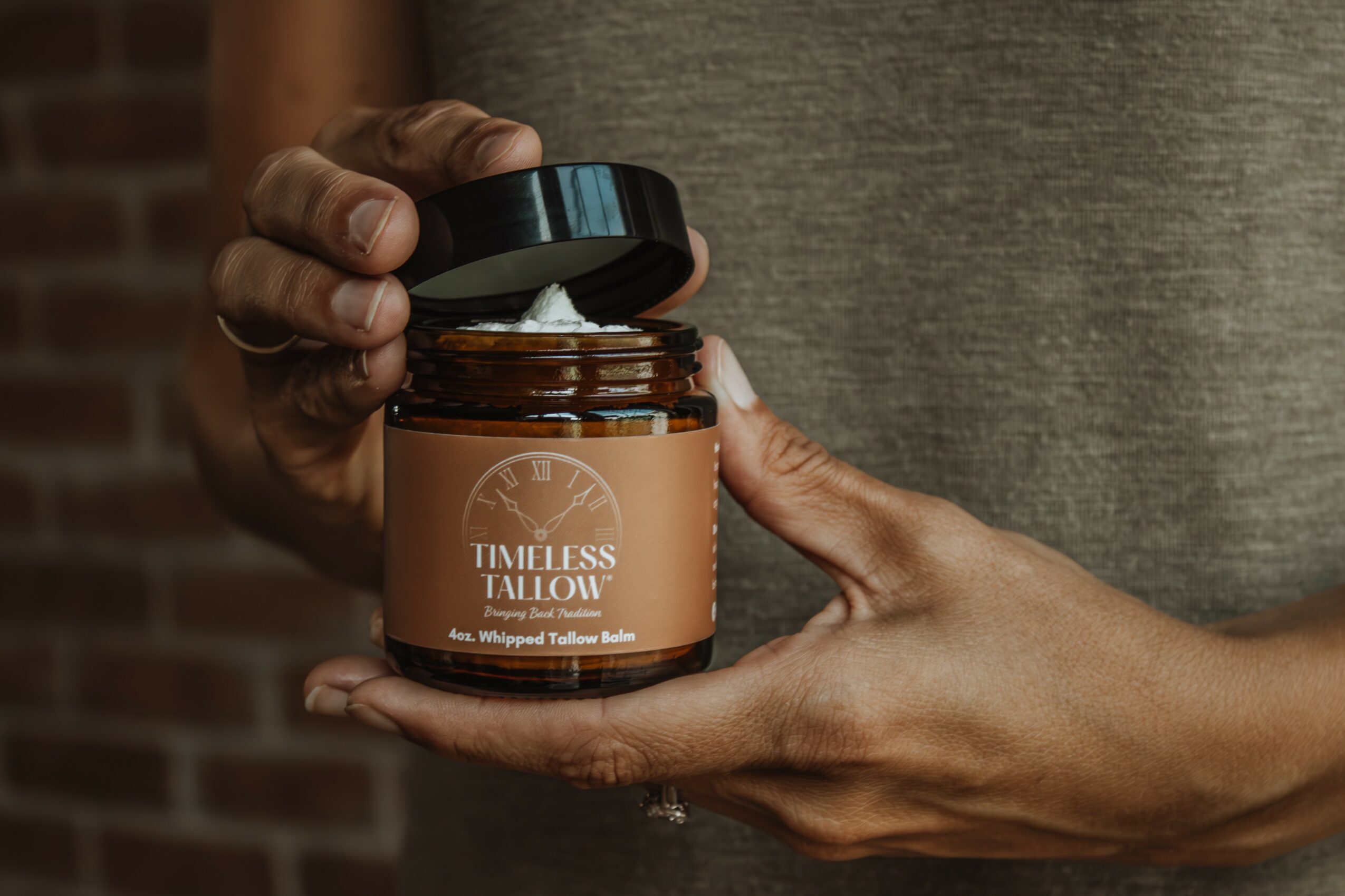 Person holding open jar labeled 'Timeless Tailor' with a black lid, in front of a brick wall, close-up view.