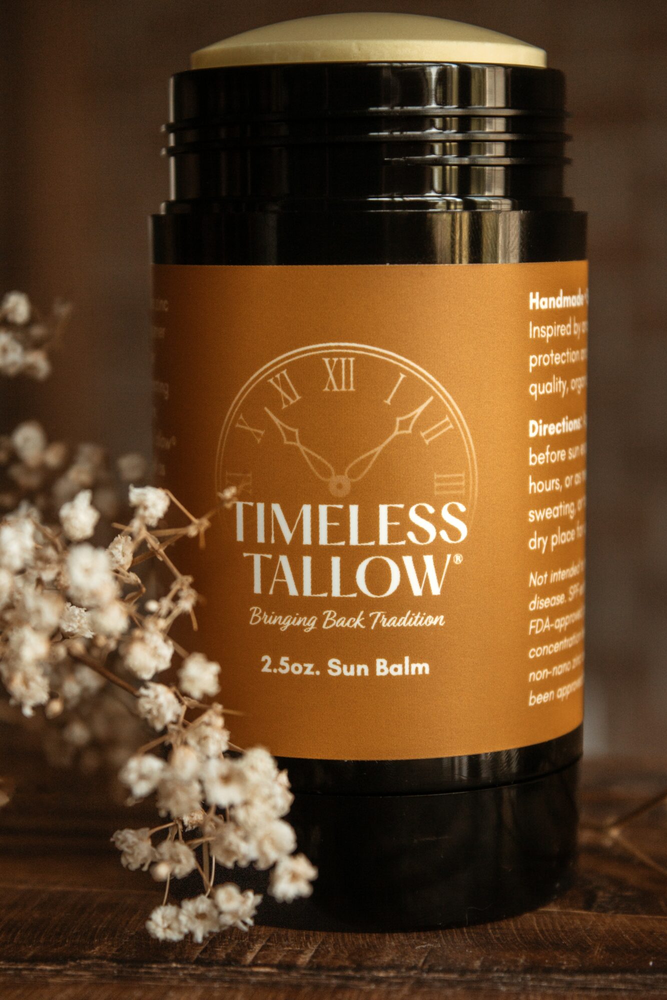 Black jar with orange label on wooden surface, with white flowers nearby, label reads 'Timeless Tallow' and '2.5 oz. Sun Balm'.