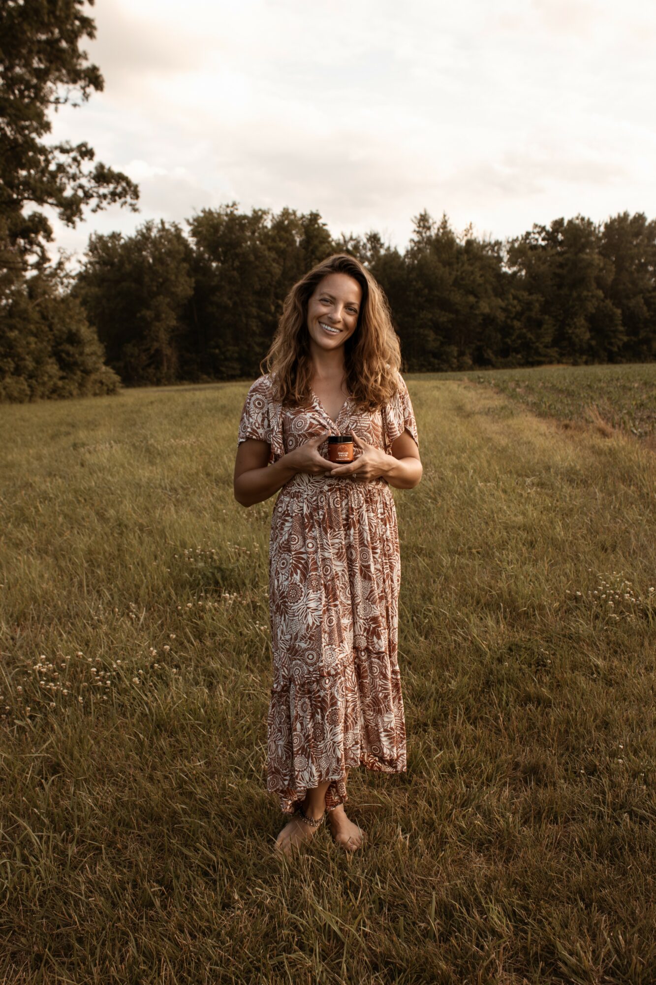 Woman standing in a grassy field holding a phone, trees in the background, cloudy sky, smiling, wearing a patterned dress.