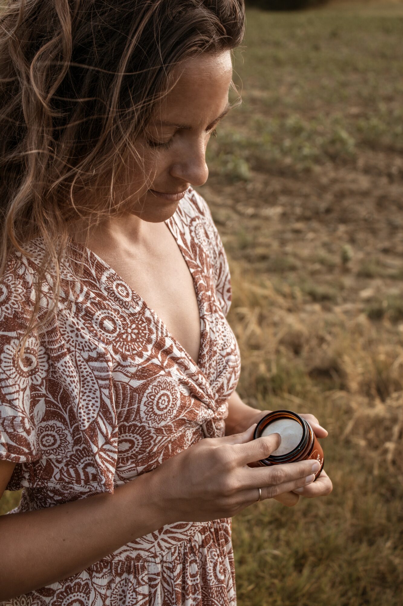 Woman with curly hair looking at a small object in her hands outdoors, wearing a patterned dress.